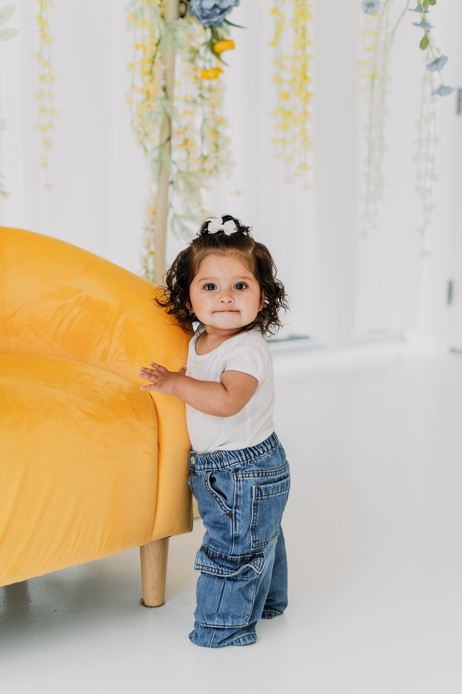 Toddler girl in jeans and white tee standing by yellow chair with floral backdrop.