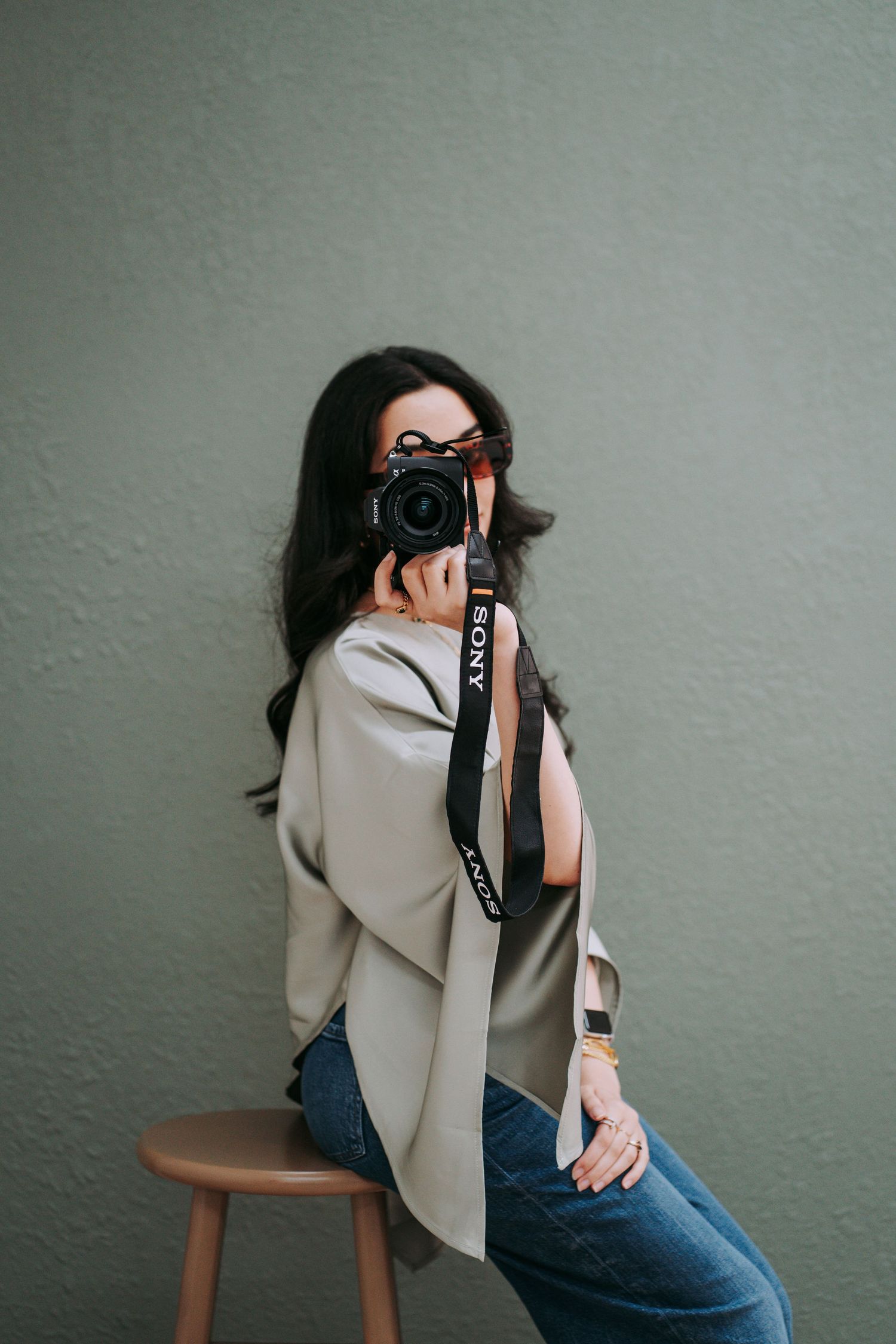 Woman photographer holding Sony camera up to her face while sitting on a stool against a green wall.