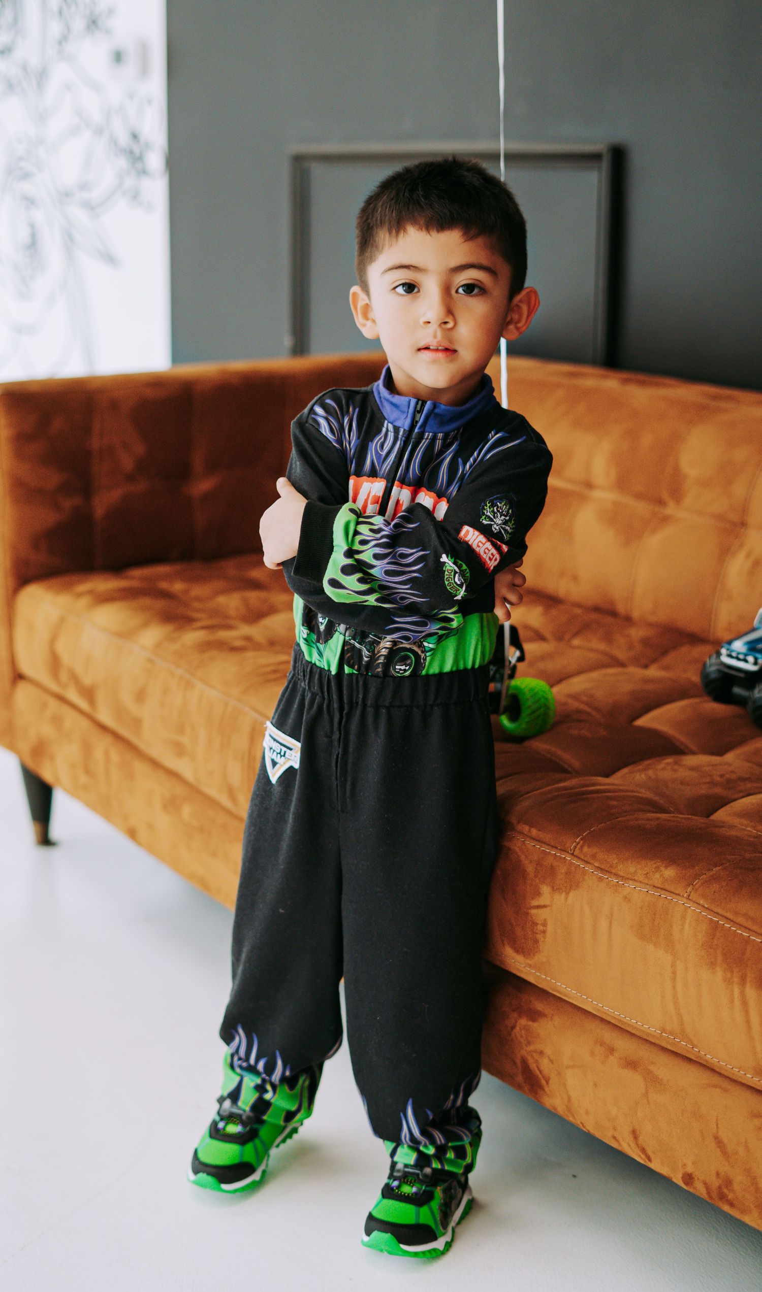 Young boy in monster truck themed outfit posing confidently by orange velvet sofa.