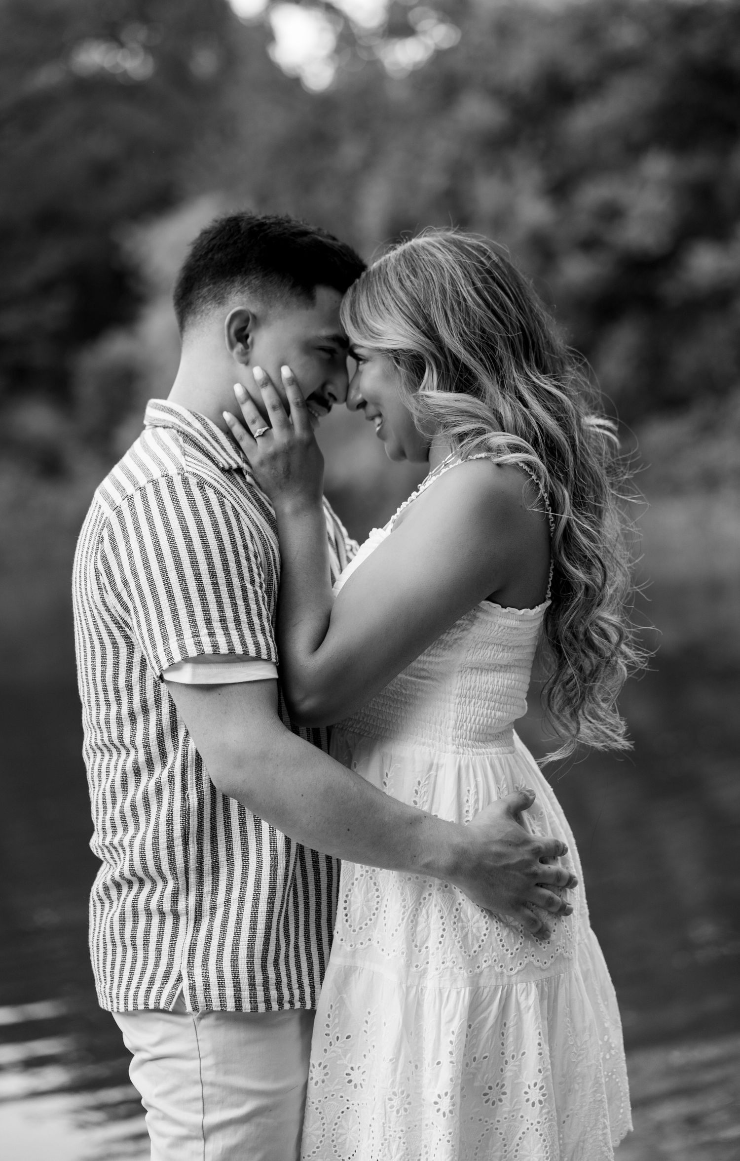 Black and white image of an engaged couple embracing near a reflective body of water outdoors.