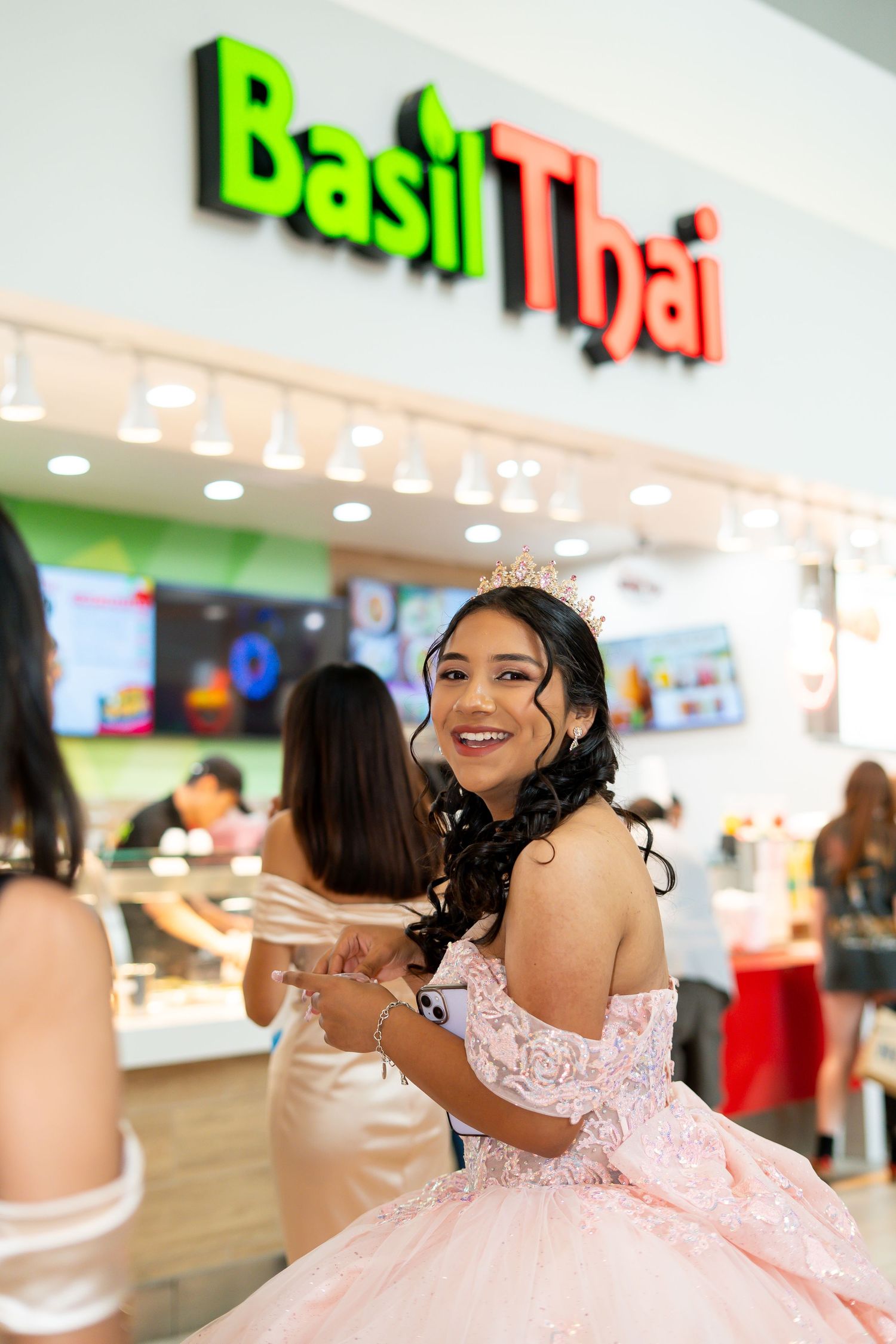 Smiling quinceañera in pink ball gown and tiara posing in front of Basil Thai restaurant in a mall.