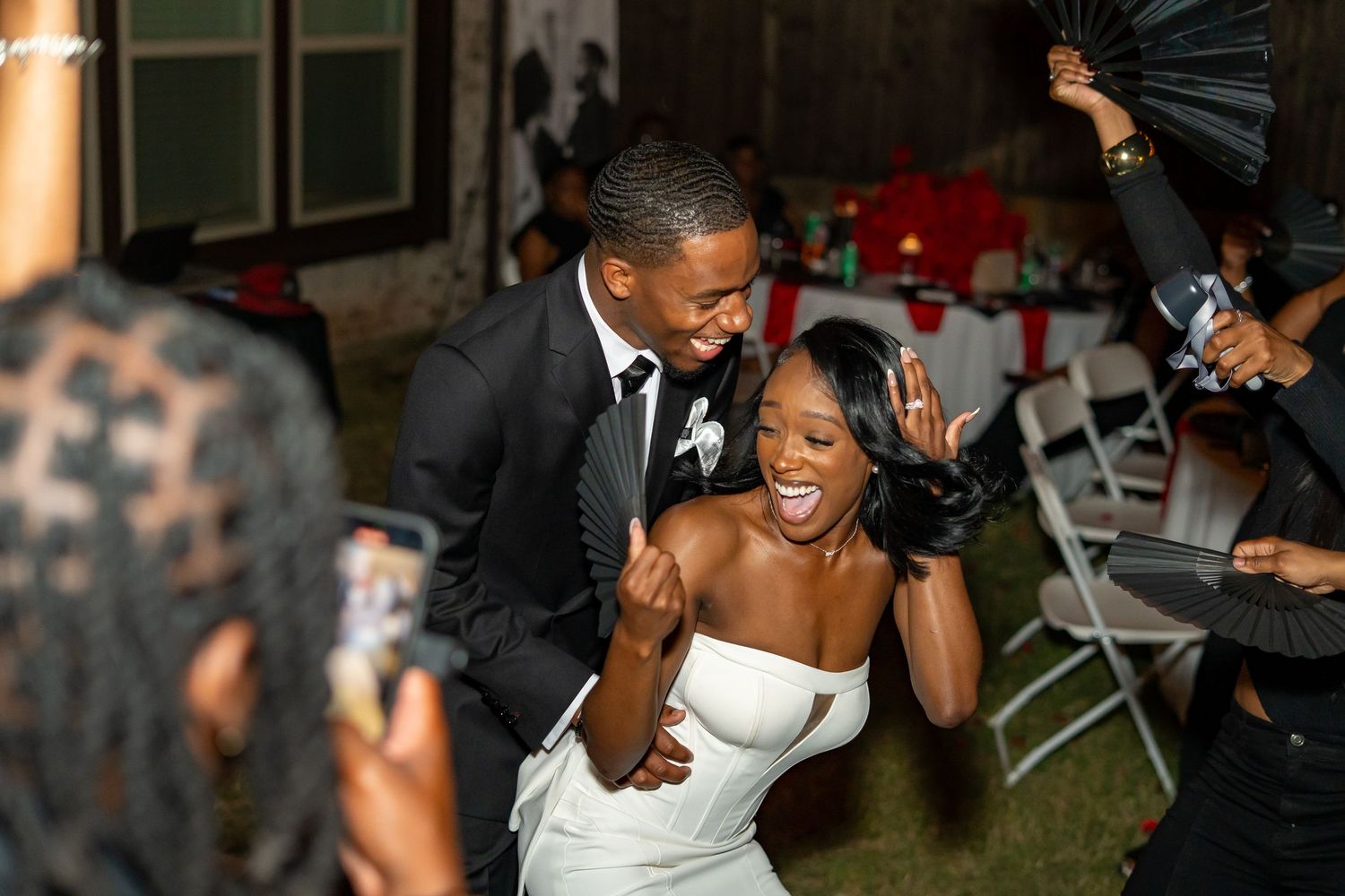Bride in white gown and groom in black suit dance joyfully at outdoor wedding reception.