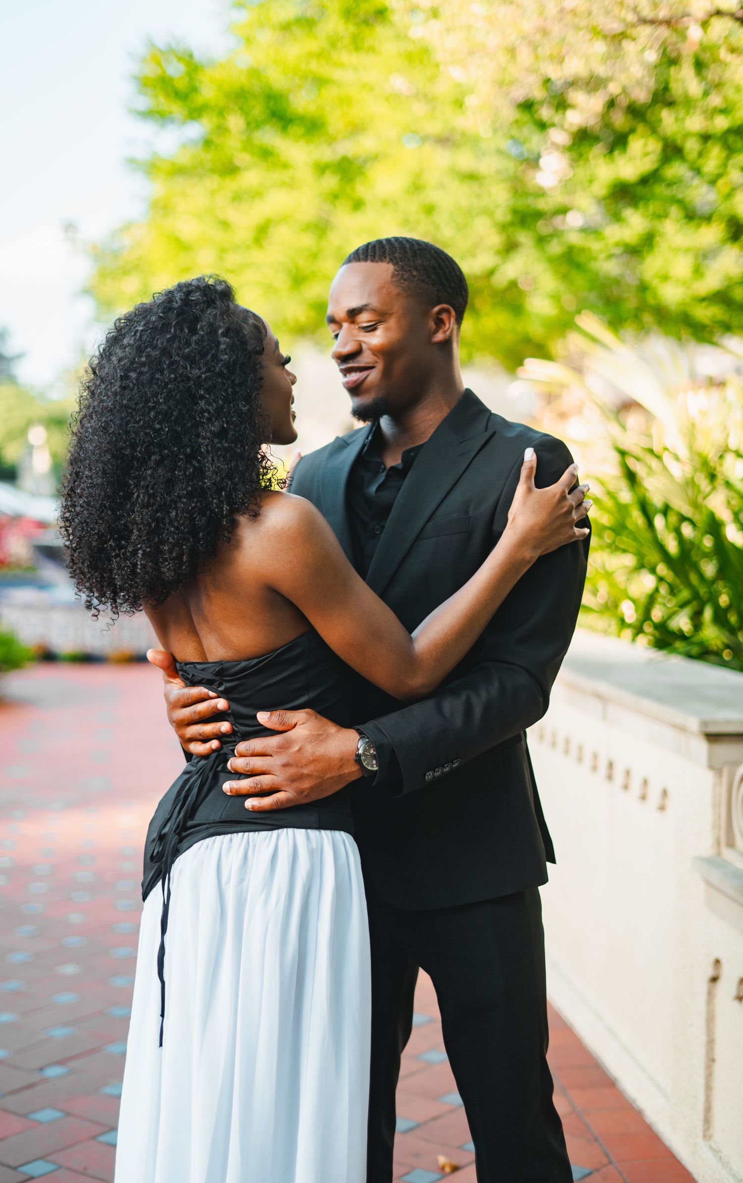 Black couple in black formal wear sharing a romantic dance embrace on a sunlit brick pathway.