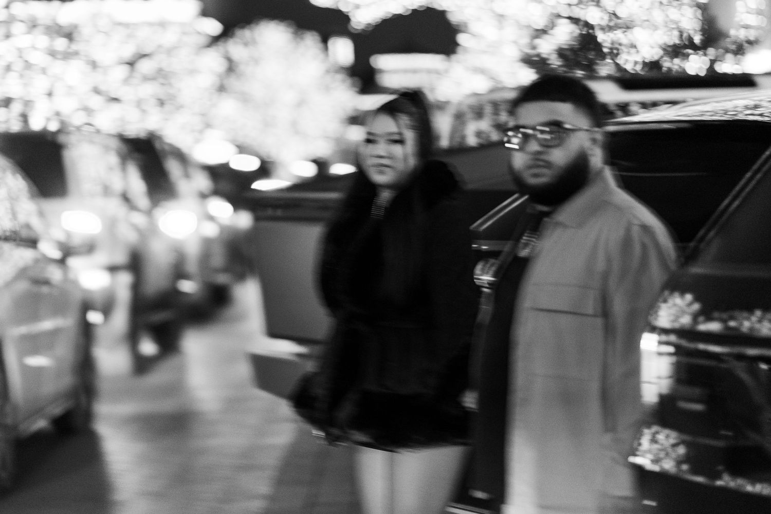 Couple walking together at night along a street adorned with festive holiday lights in black and white.