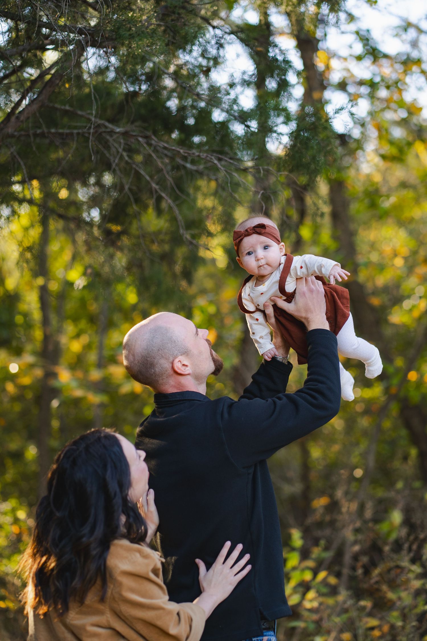 Parents lifting a baby girl in the air outdoors surrounded by colorful autumn foliage in a joyful moment.