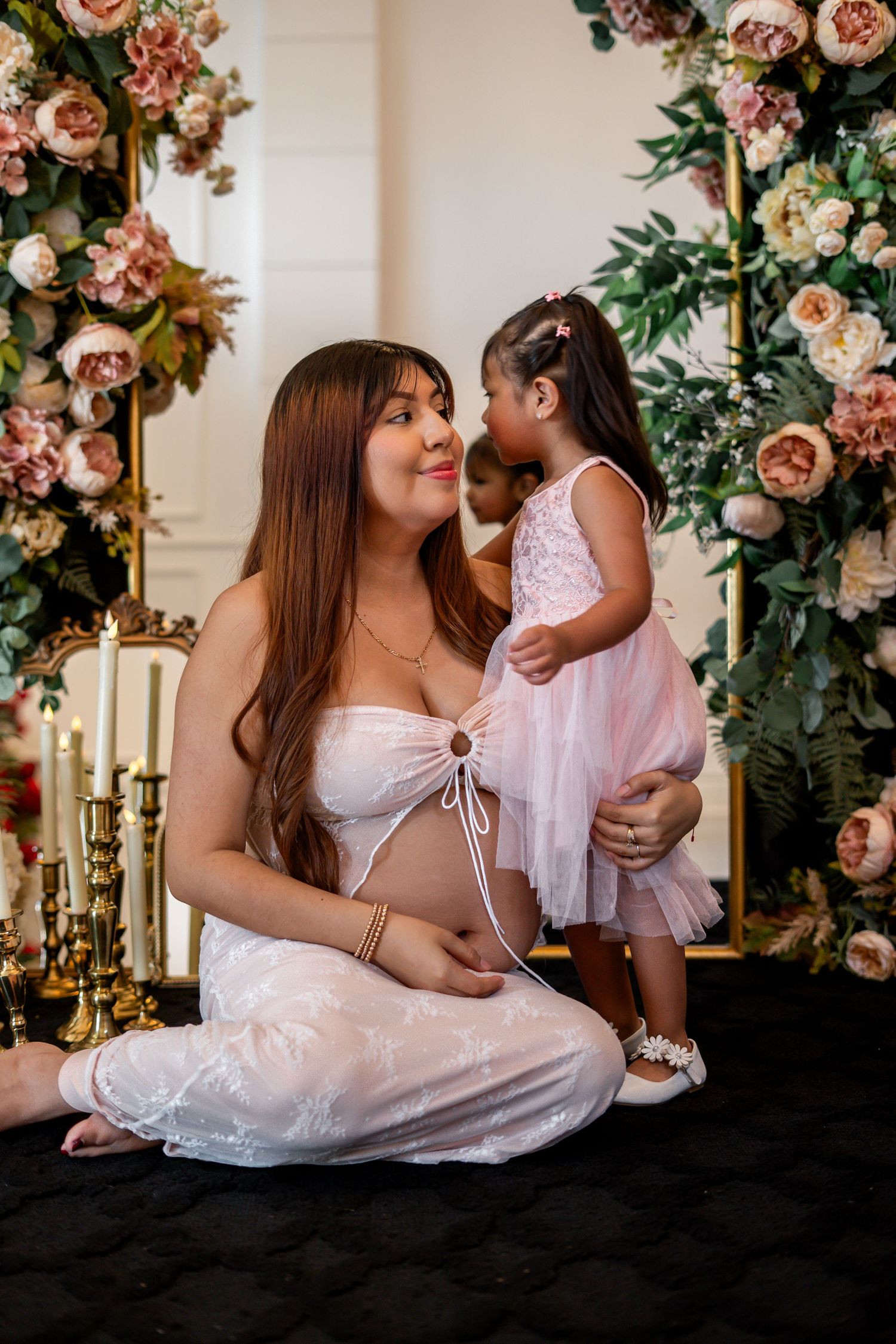 Pregnant woman in white lace outfit sitting with a toddler girl in pink dress before a floral arch.