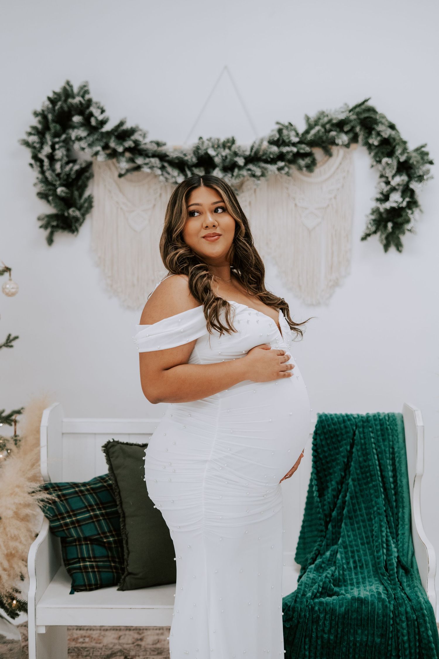 Maternity photo of woman in white fitted dress beside white bench with holiday greenery and macrame backdrop.