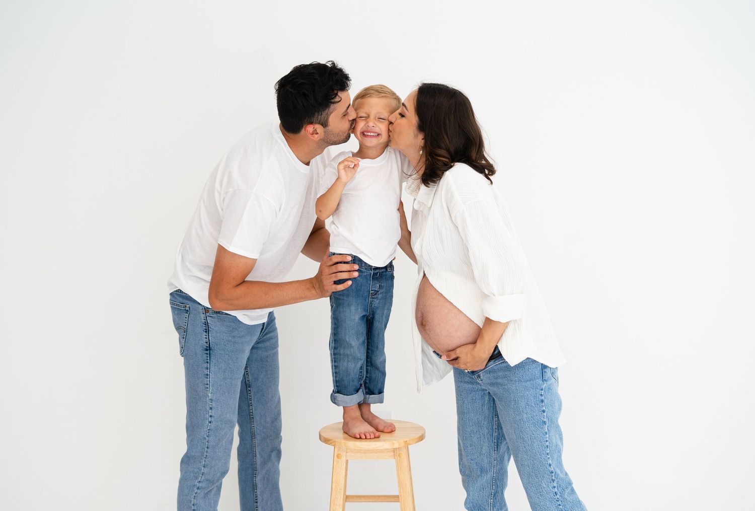 Couple in white shirts and jeans kissing smiling toddler on stool during maternity photoshoot.