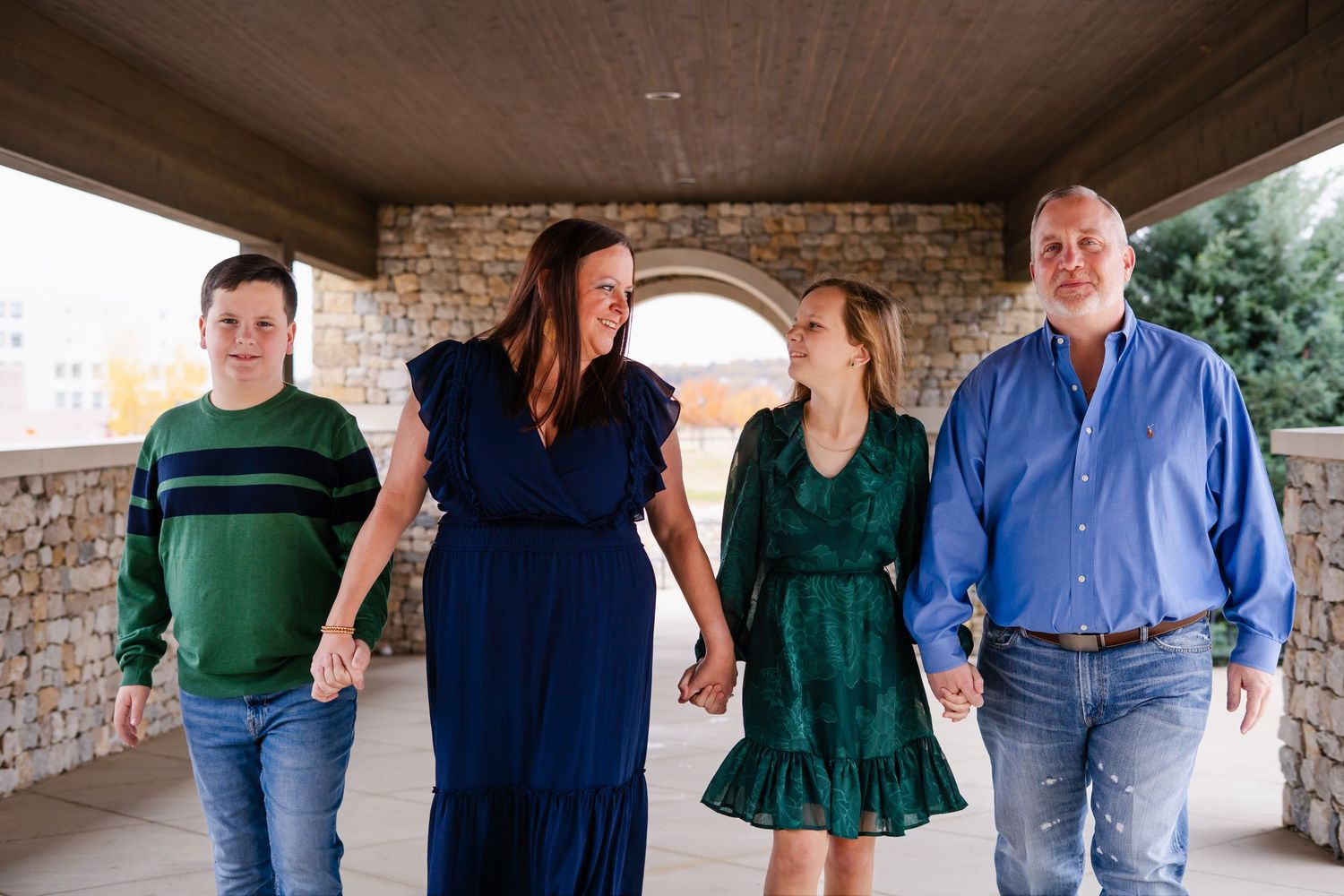 A family of four holds hands and walks together under a covered stone pavilion while smiling.