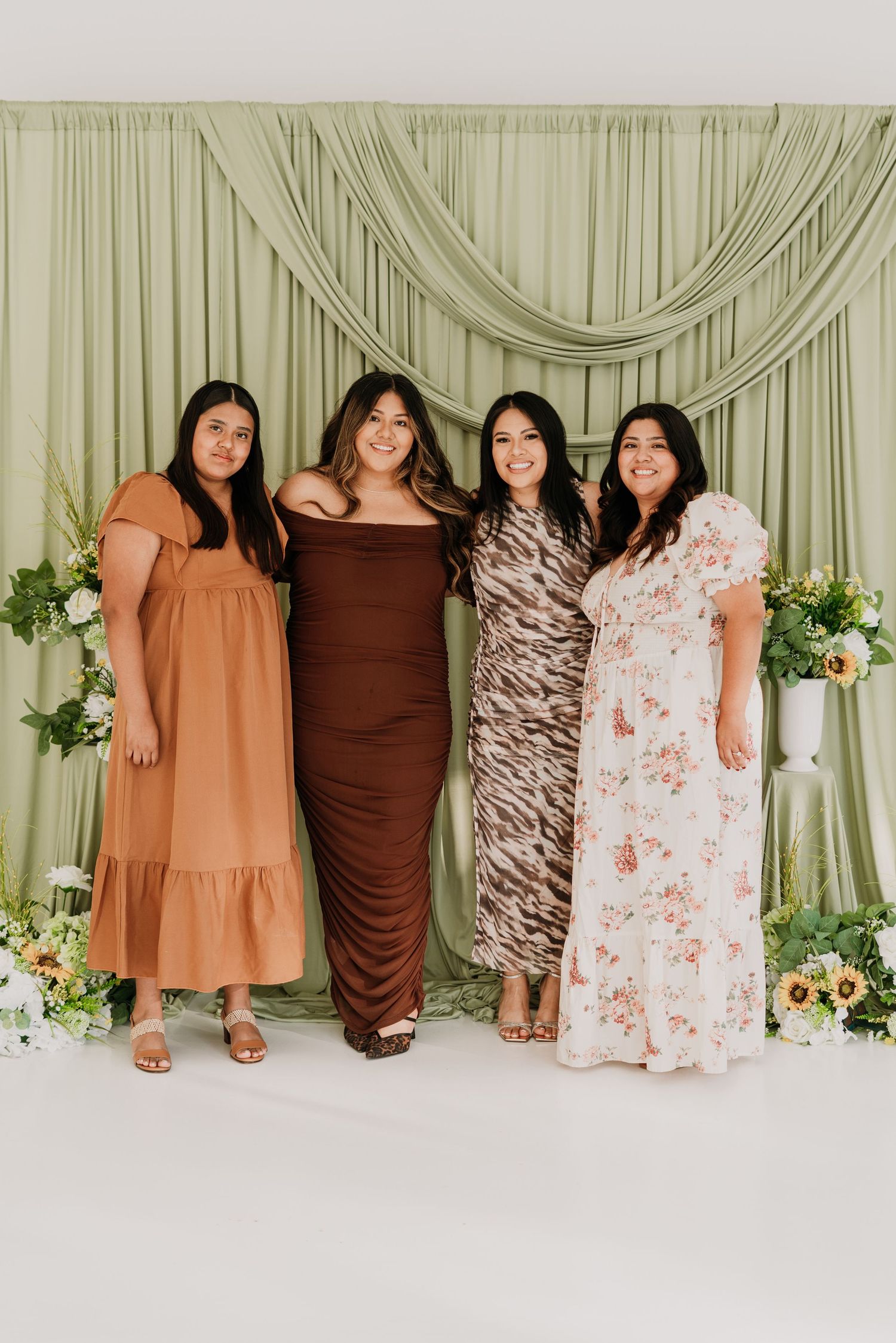 Four women in elegant maxi dresses pose together in front of a draped green backdrop with floral arrangements.