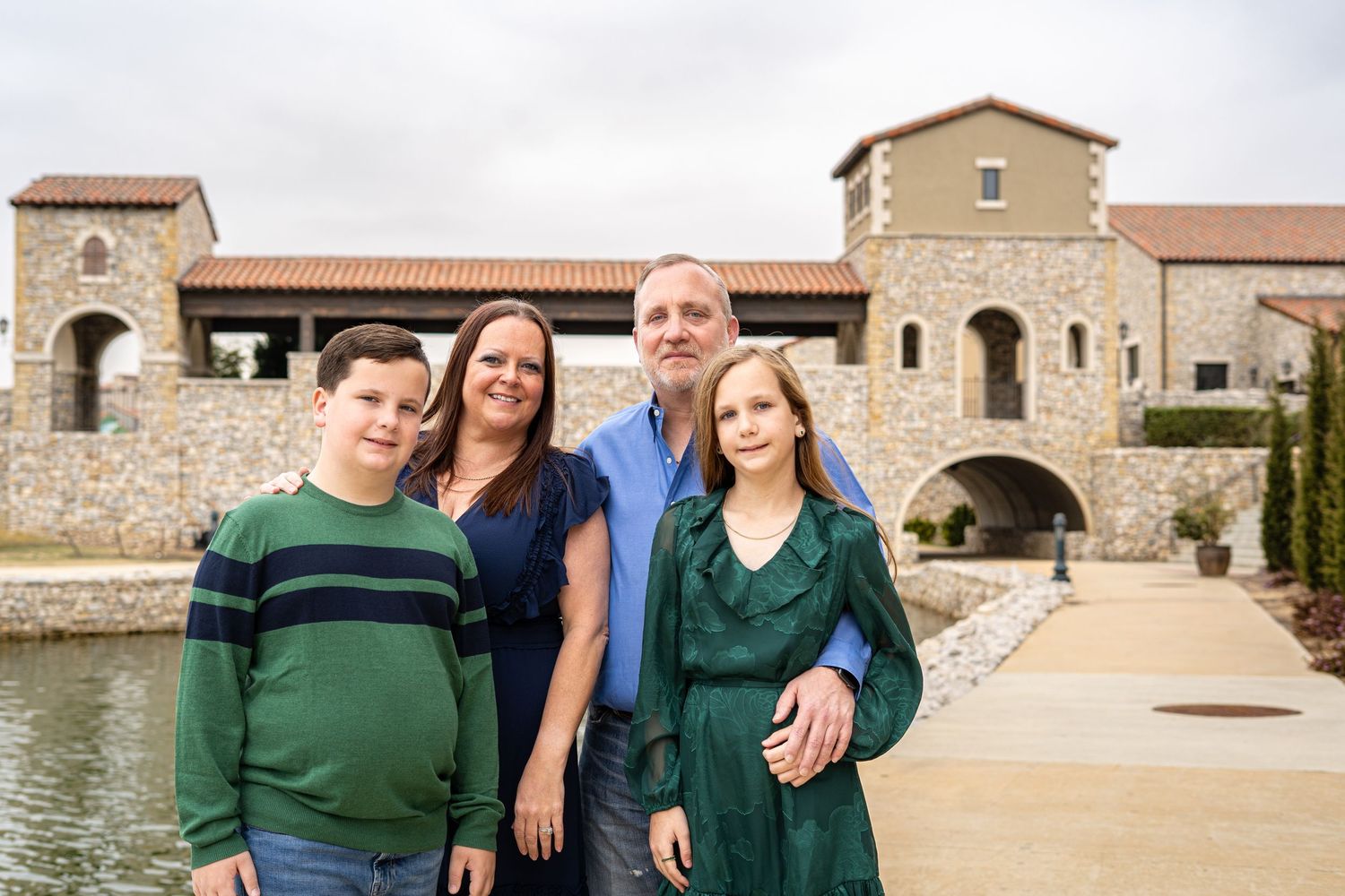 A family of four stands together near a beautiful stone building reflected in a calm waterfront.