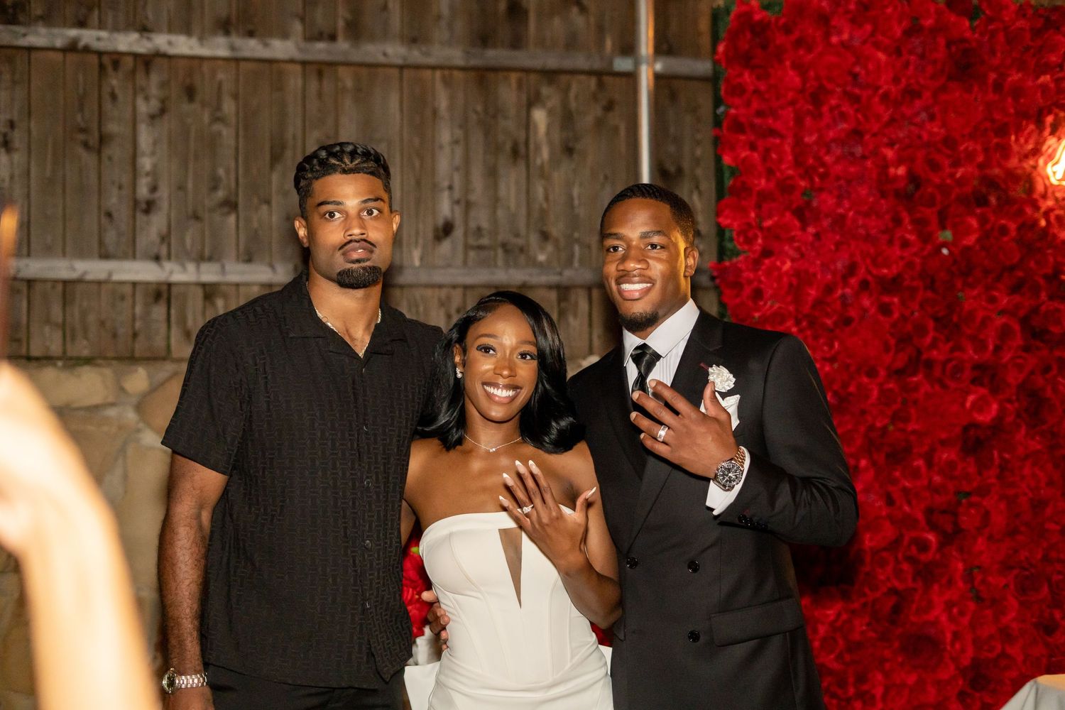 Bride and groom posing with a guest in front of a stunning red rose floral wall backdrop.