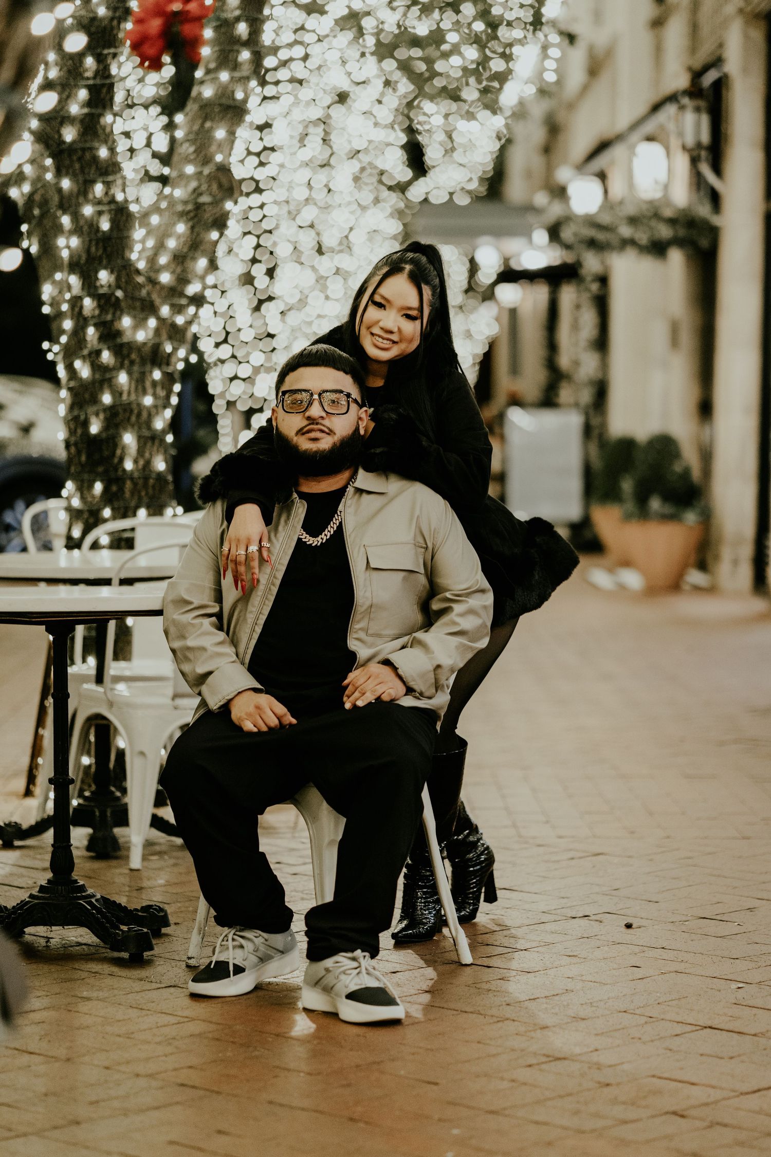 A couple poses together on a festive lit outdoor patio, man seated and woman standing behind him.