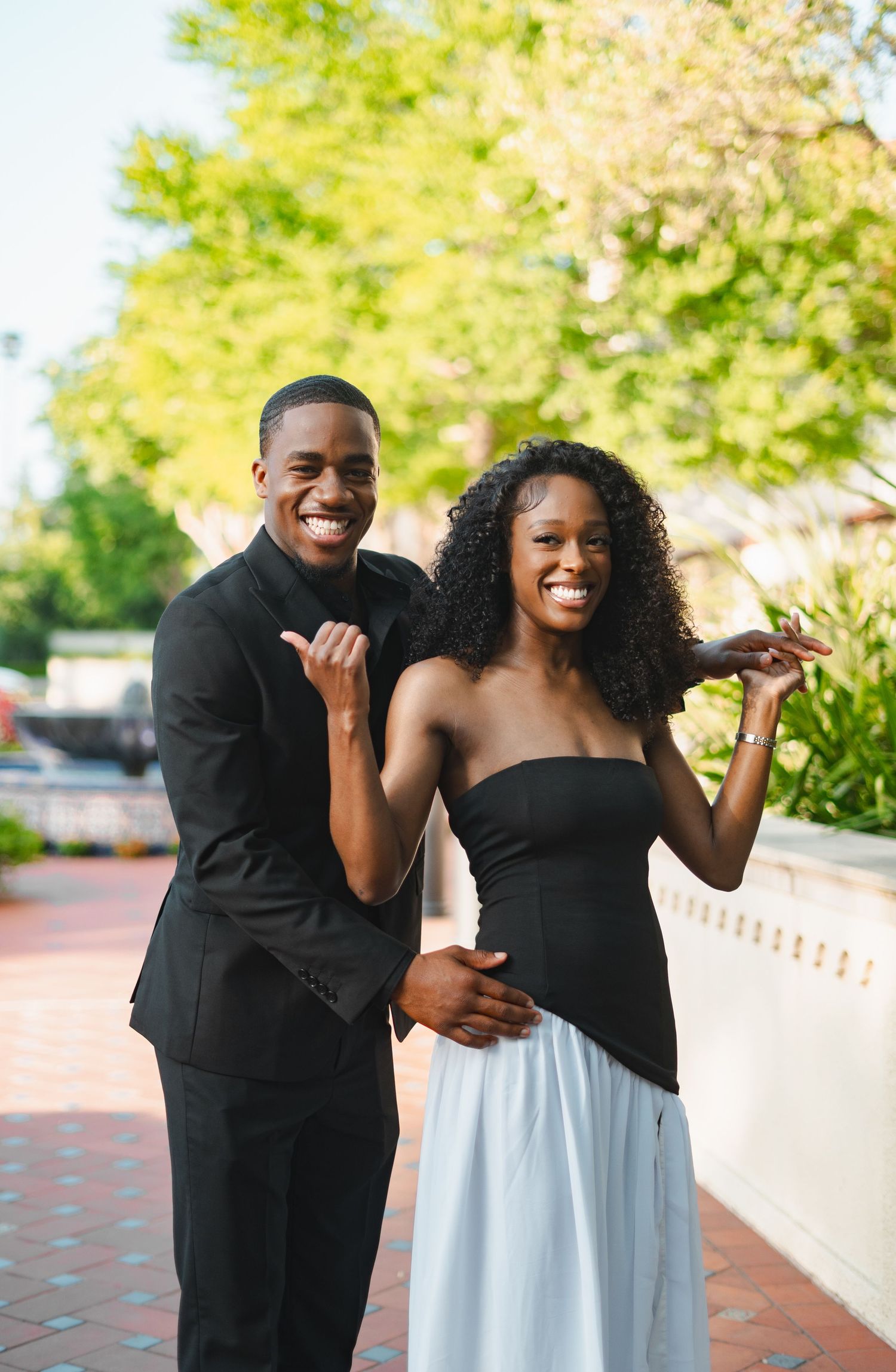 Smiling Black couple in black formal wear posing playfully together on a brick pathway outdoors.