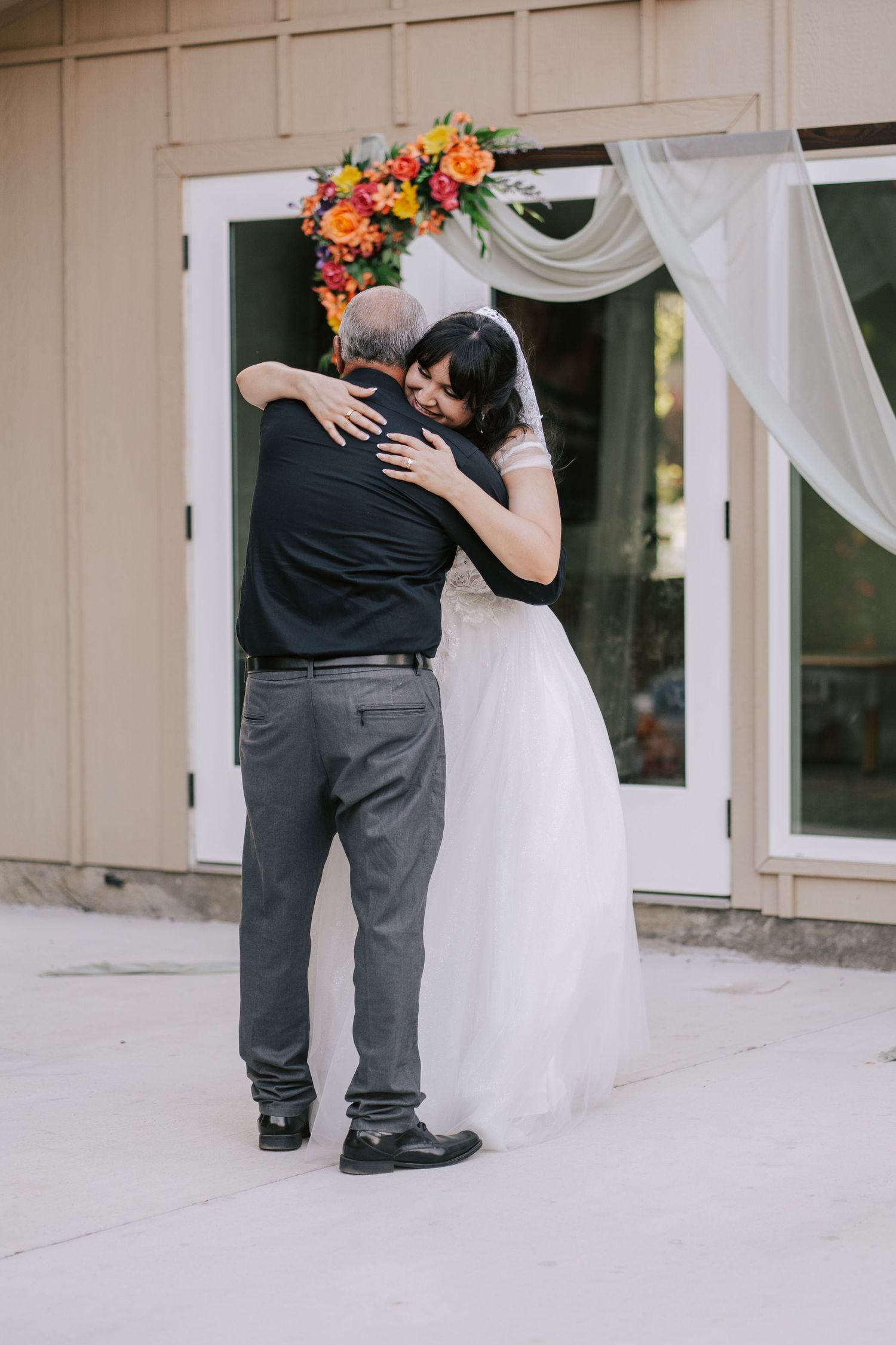 Bride in white gown shares emotional hug with father near colorful floral wedding arch.
