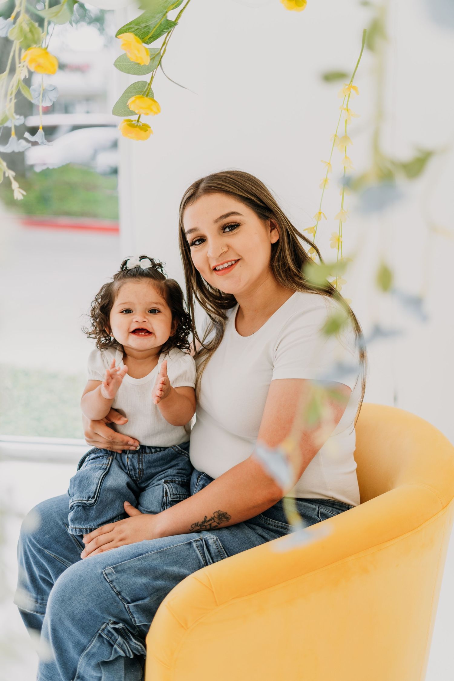 A young woman and toddler girl in matching white tops and jeans sit together on a yellow chair with floral decor.