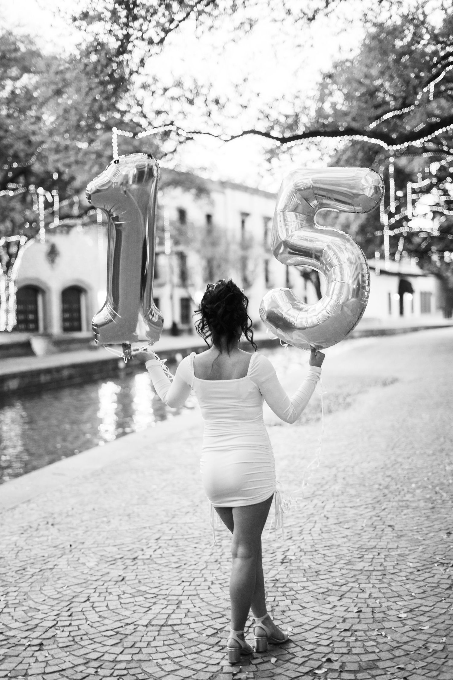 Black and white photo of quinceañera girl holding large number 15 balloons by a scenic waterway.