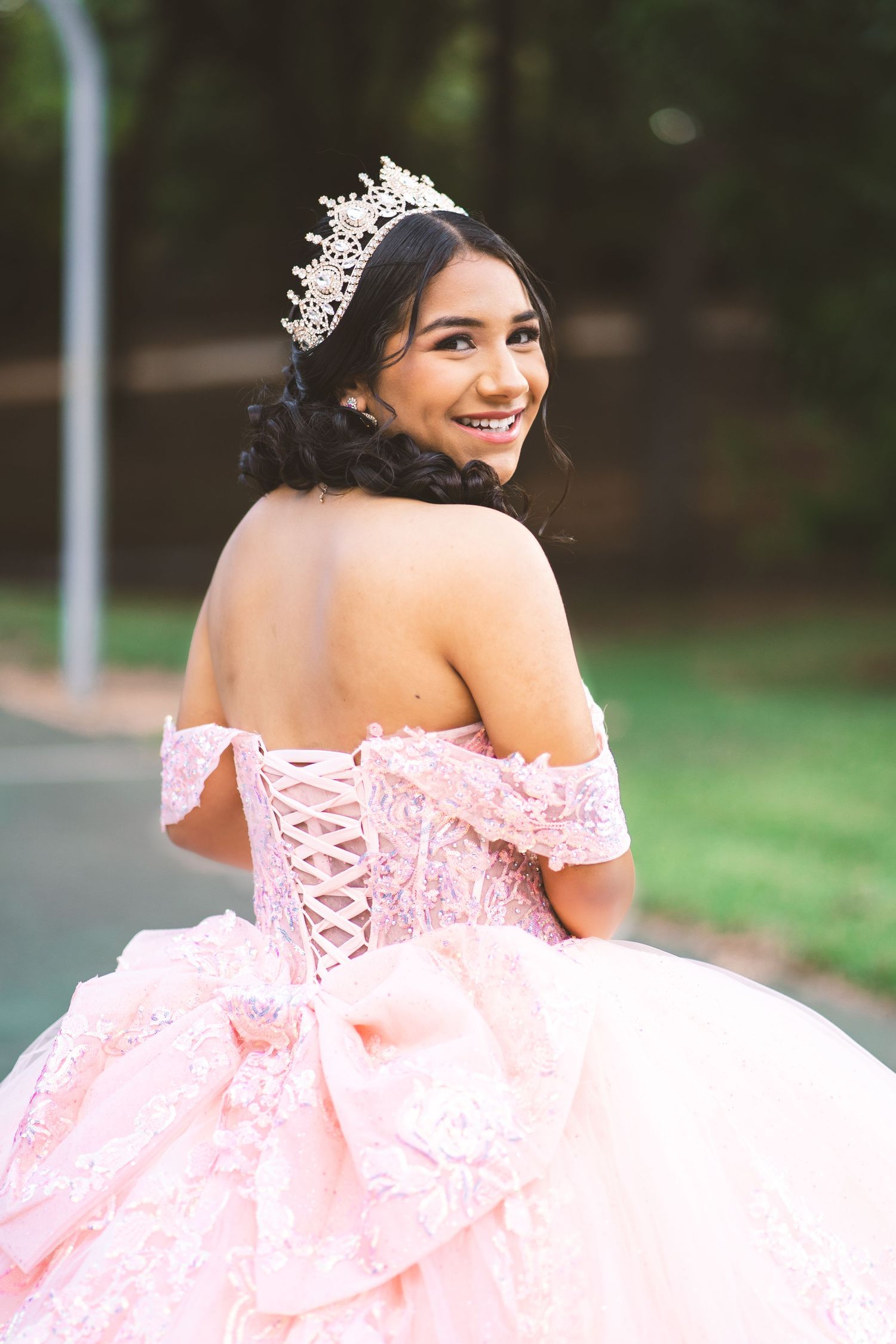Young woman in a pink quinceañera ball gown with lace corset back and crystal tiara, smiling outdoors.
