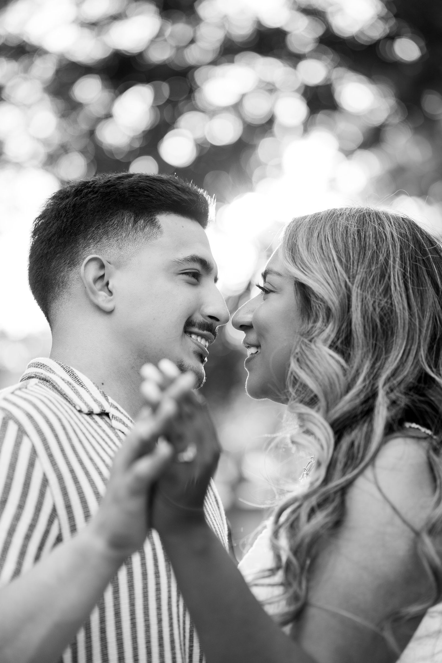 Black and white photo of a smiling couple touching noses and holding hands outdoors.