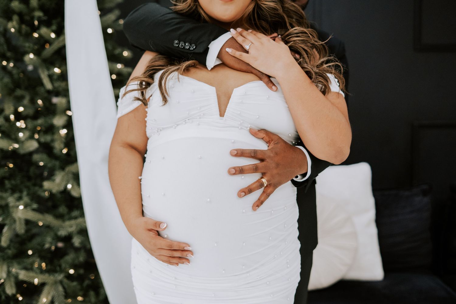 Close-up of a pregnant woman in a white dress with her partner embracing her baby bump near a Christmas tree.