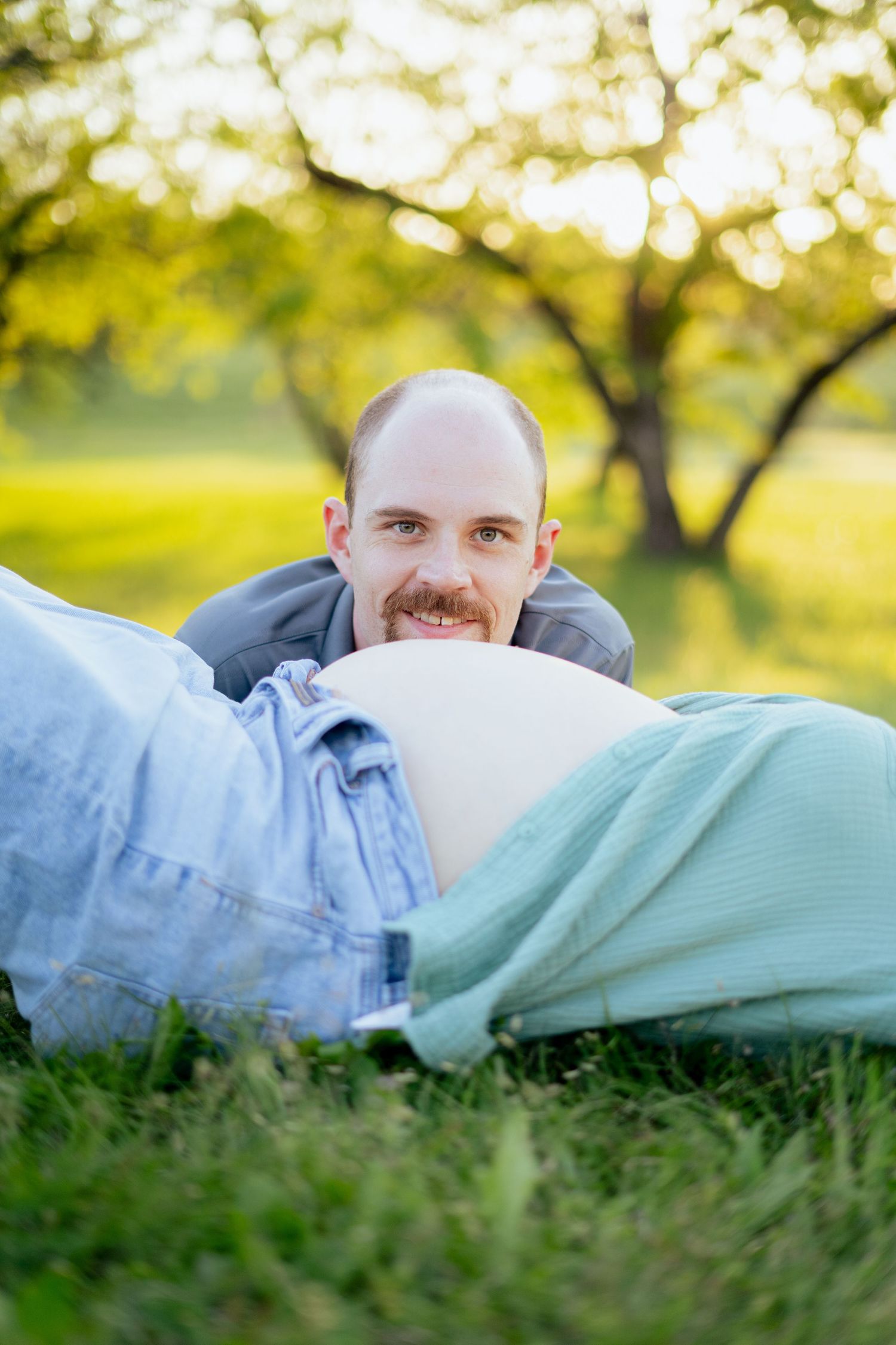 Smiling man with mustache rests chin near pregnant woman's bare belly while lying on grass in sunny park.