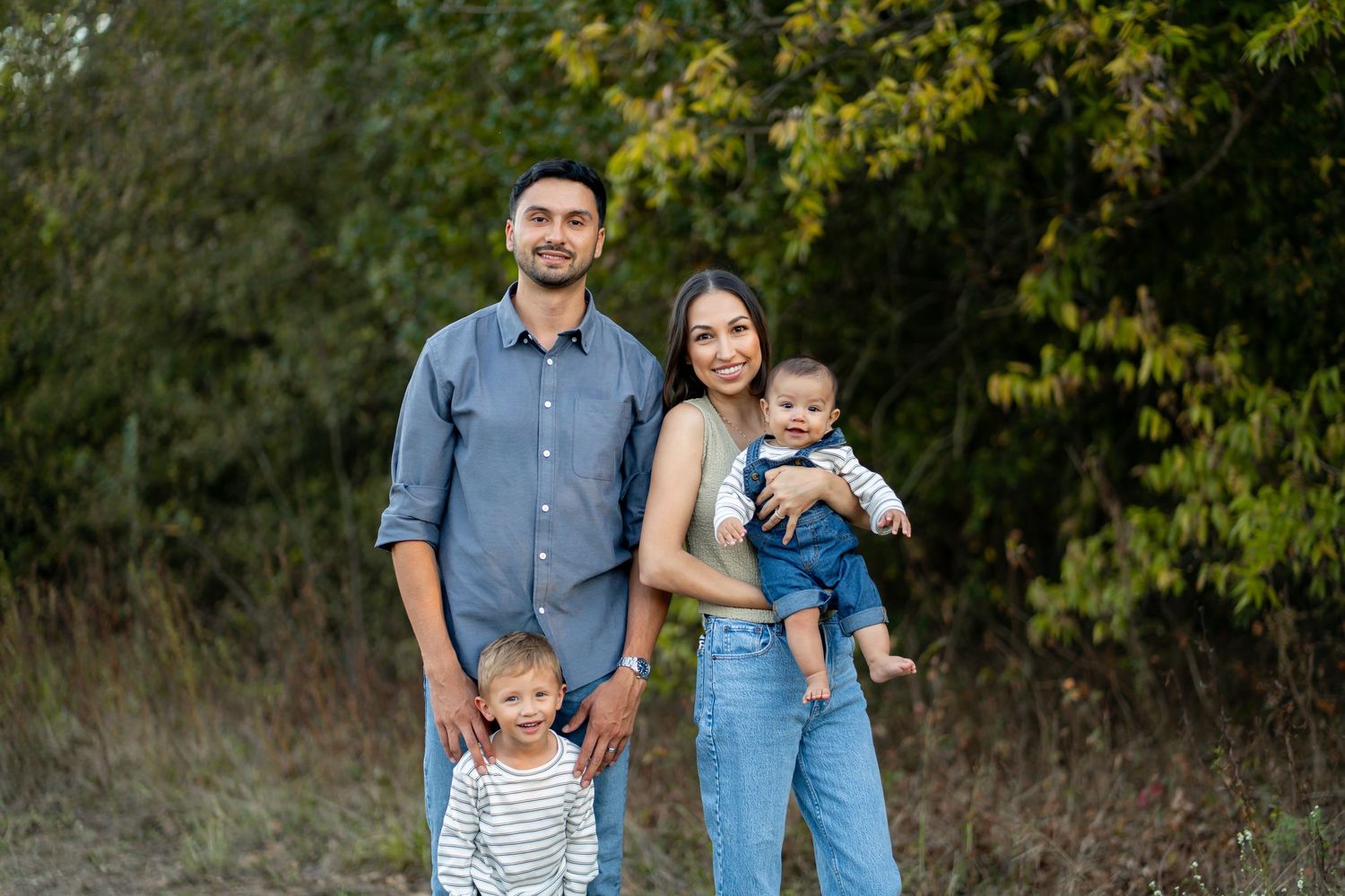 Young family of four smiling together outdoors in front of lush green autumn foliage.