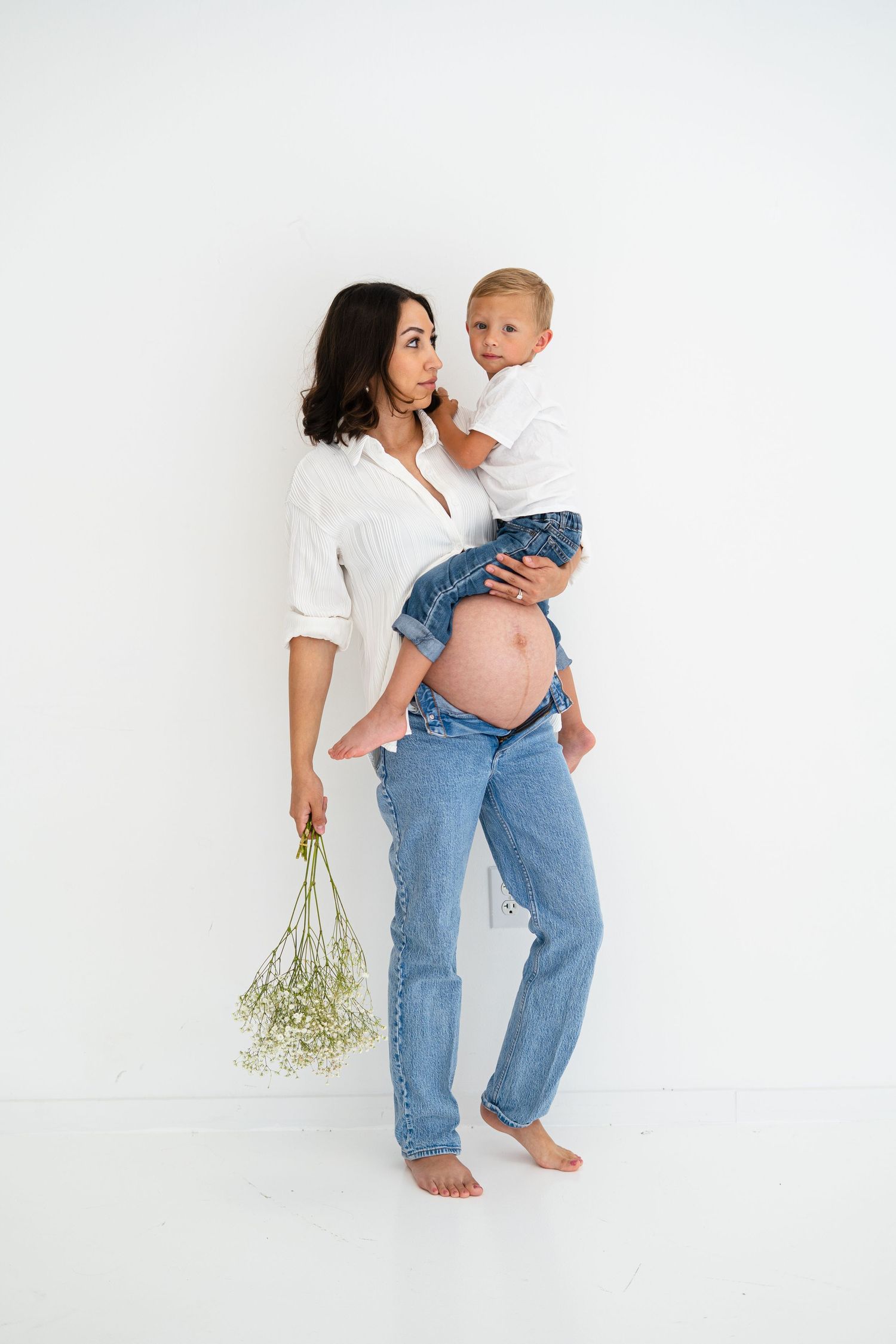 A pregnant woman in a white shirt holds her young son while carrying a bouquet of white flowers.