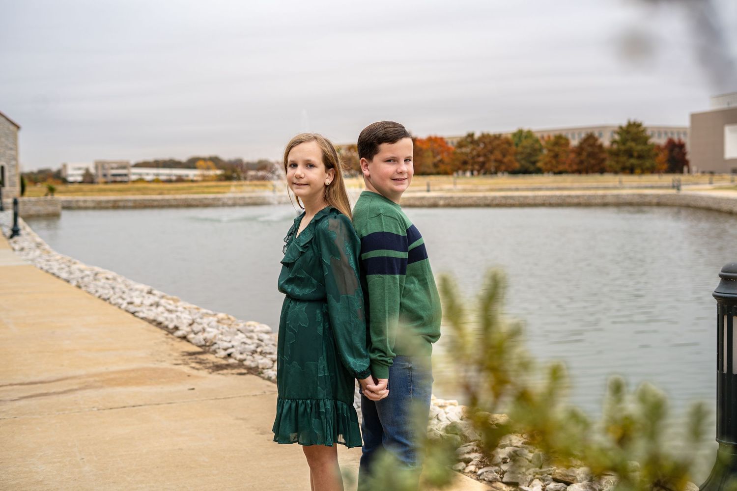 A boy and girl in coordinating green outfits stand back to back near a tranquil lakeside path.