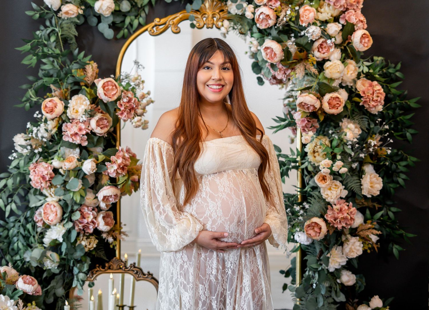 Smiling pregnant woman in white lace gown standing before a floral arch with pink roses and greenery.