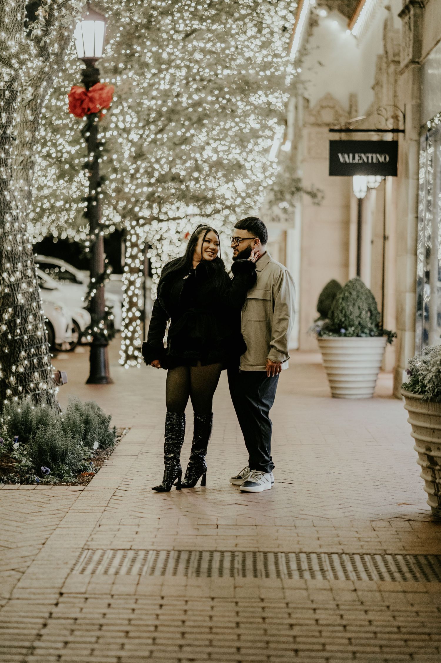 A couple walks hand in hand along a holiday-lit outdoor shopping street near Valentino store.