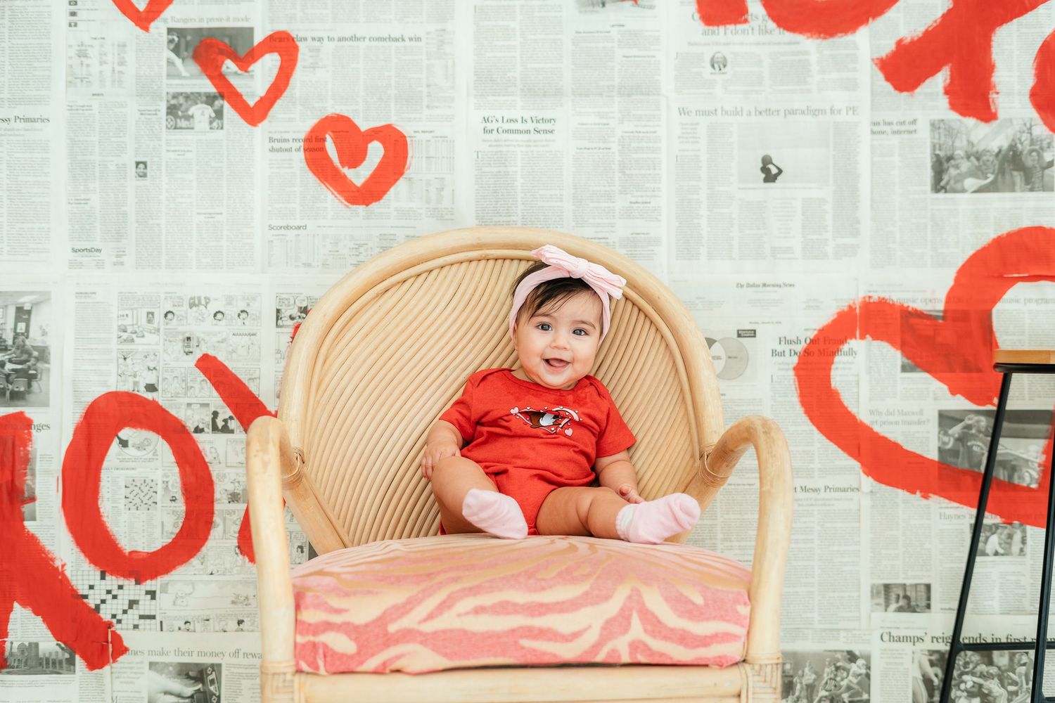 Cute baby in red outfit sits in wicker chair before Valentine's Day XOXO newspaper backdrop.