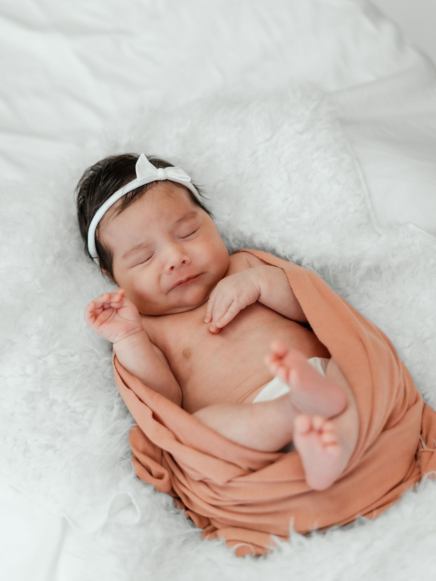 Sleeping newborn baby girl wrapped in peach swaddle with white bow headband on fluffy white blanket.