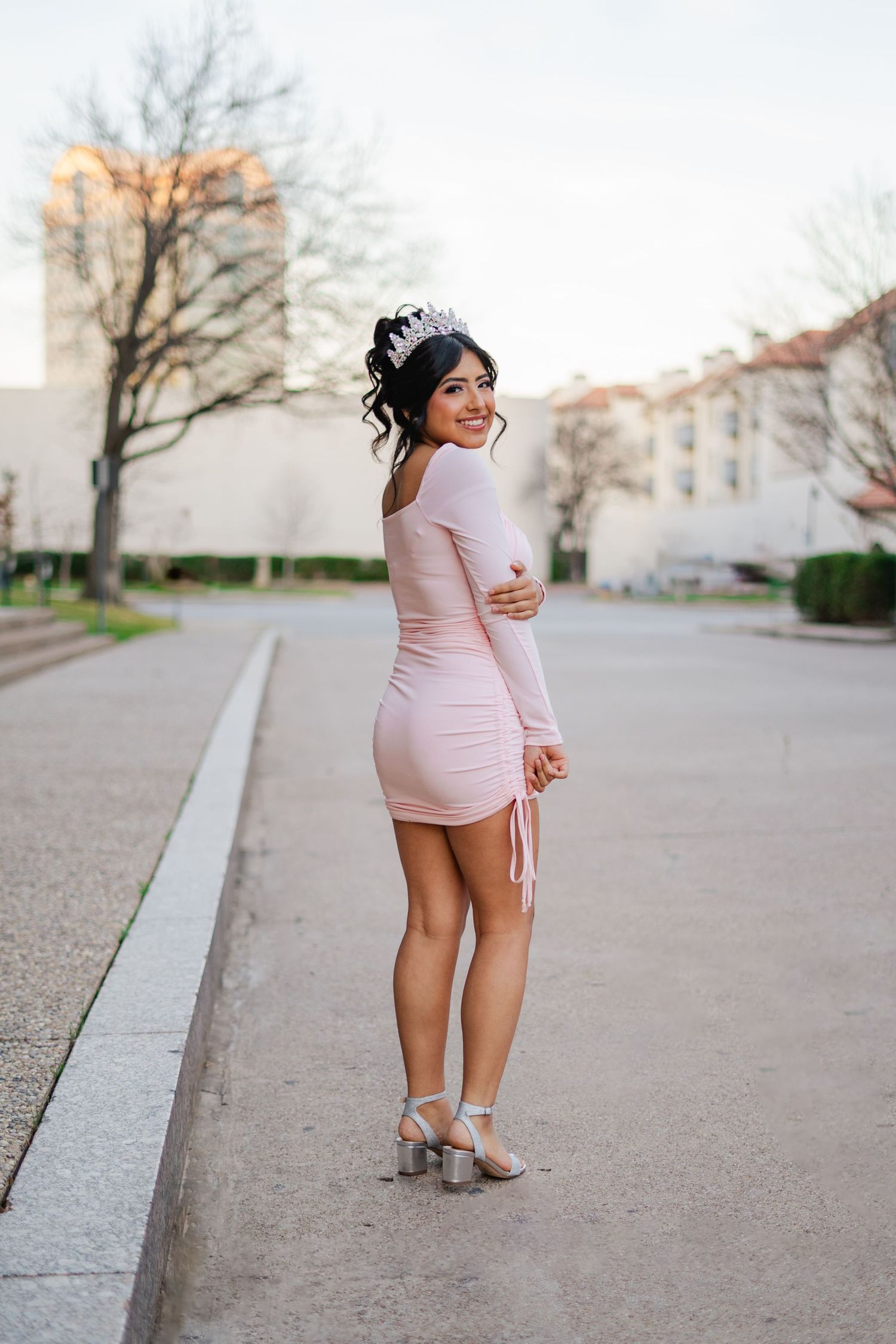 Young woman in pink bodycon dress and tiara smiles over shoulder during outdoor quinceañera photoshoot.