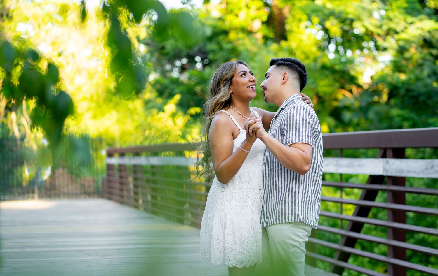 Couple holding hands and smiling at each other on a wooden bridge surrounded by lush green trees.