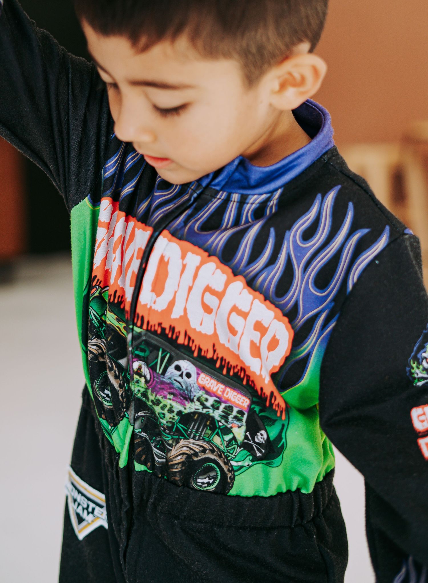 Boy in Grave Digger outfit sitting small on orange sofa surrounded by purple and green balloons.