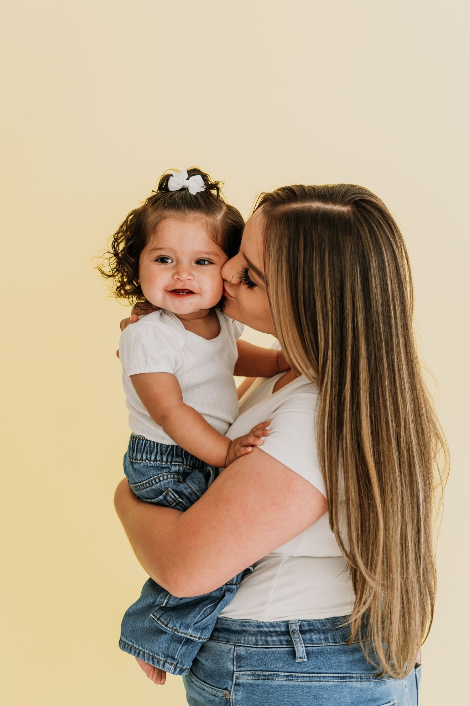 A mother with long blonde hair kisses her smiling toddler girl in a white top and jeans against a yellow background.