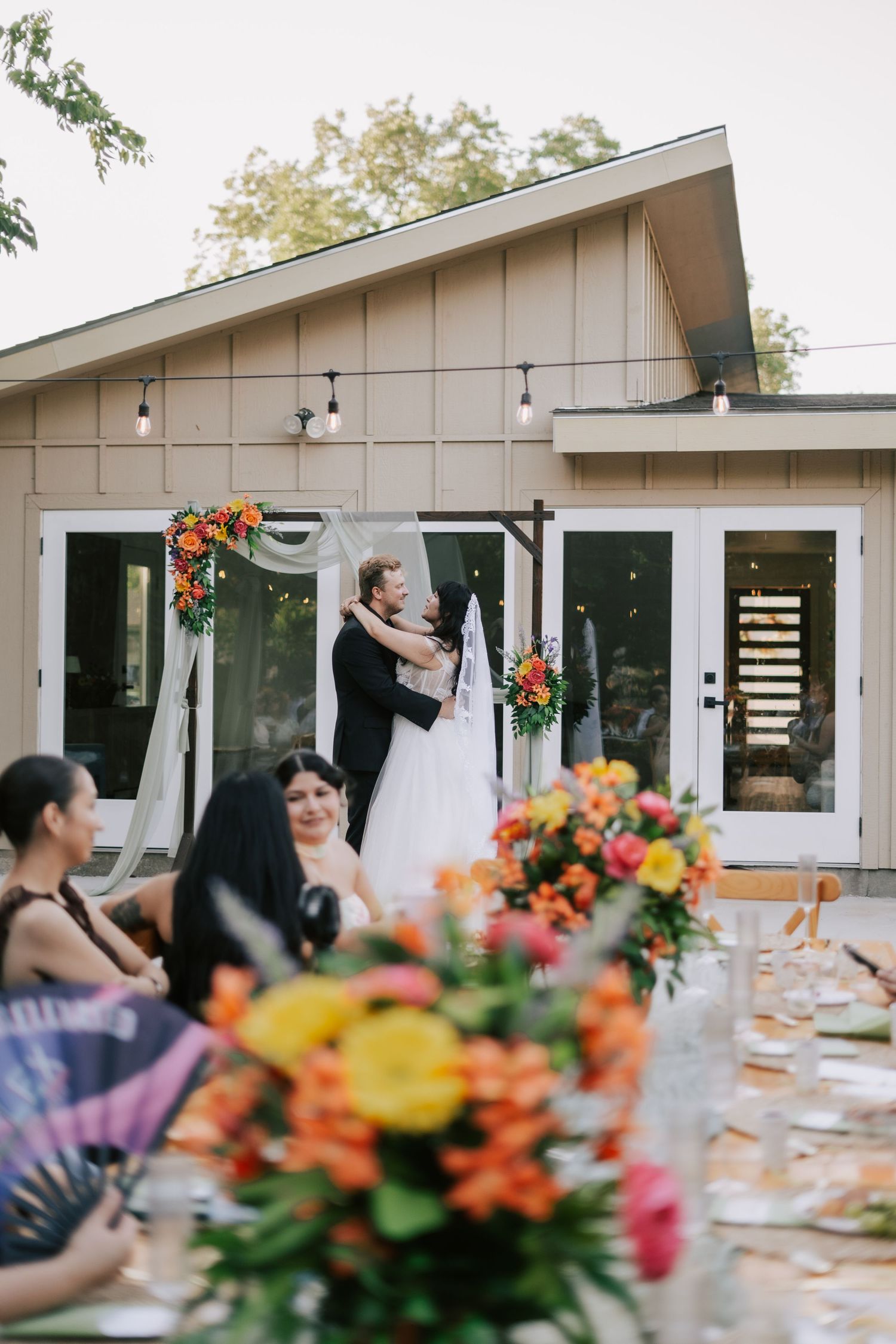 Bride and groom share first kiss at outdoor wedding ceremony with colorful floral arch decorations.