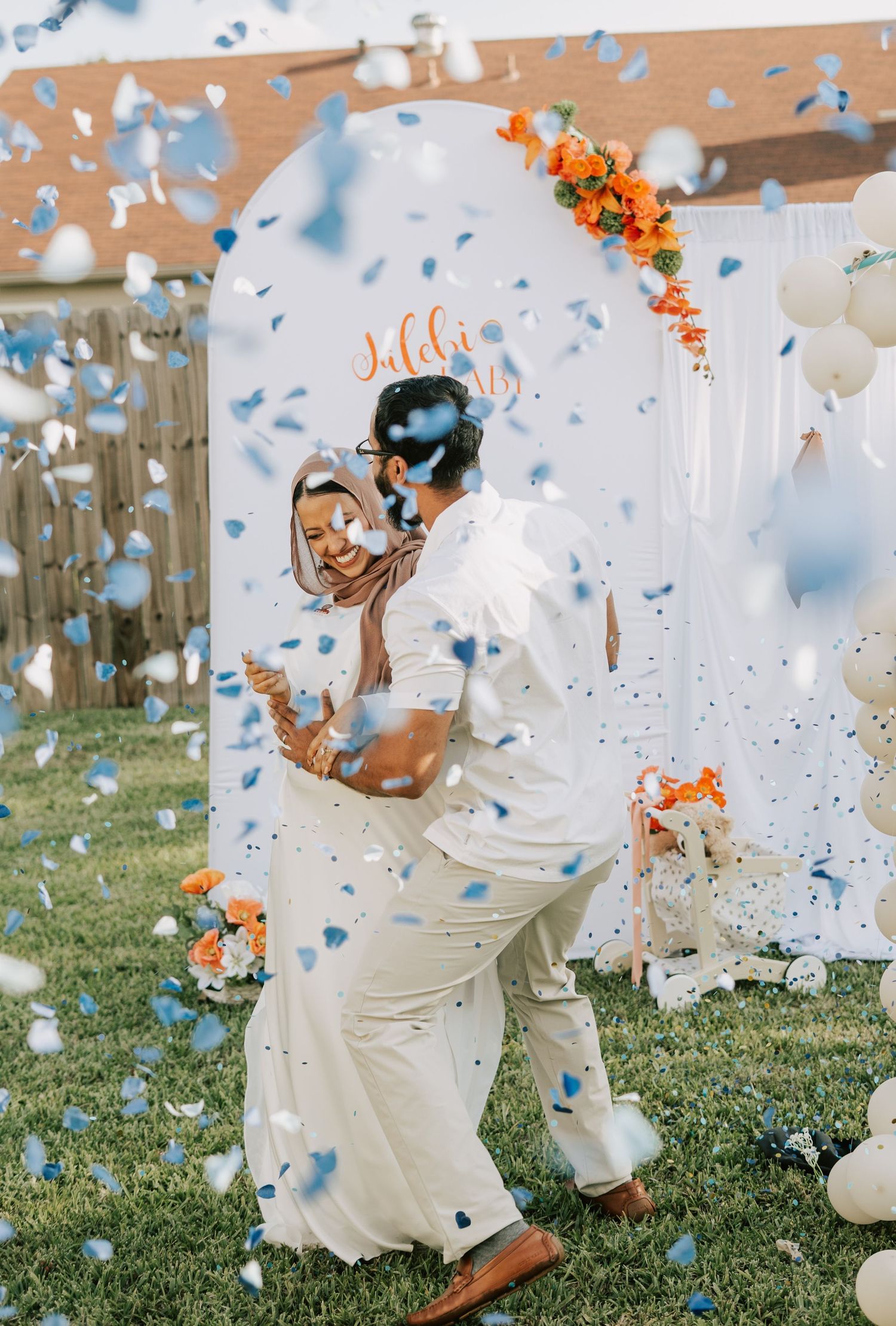 Couple dancing outdoors at a wedding celebration with blue confetti falling, white arch backdrop, and orange floral decorations.