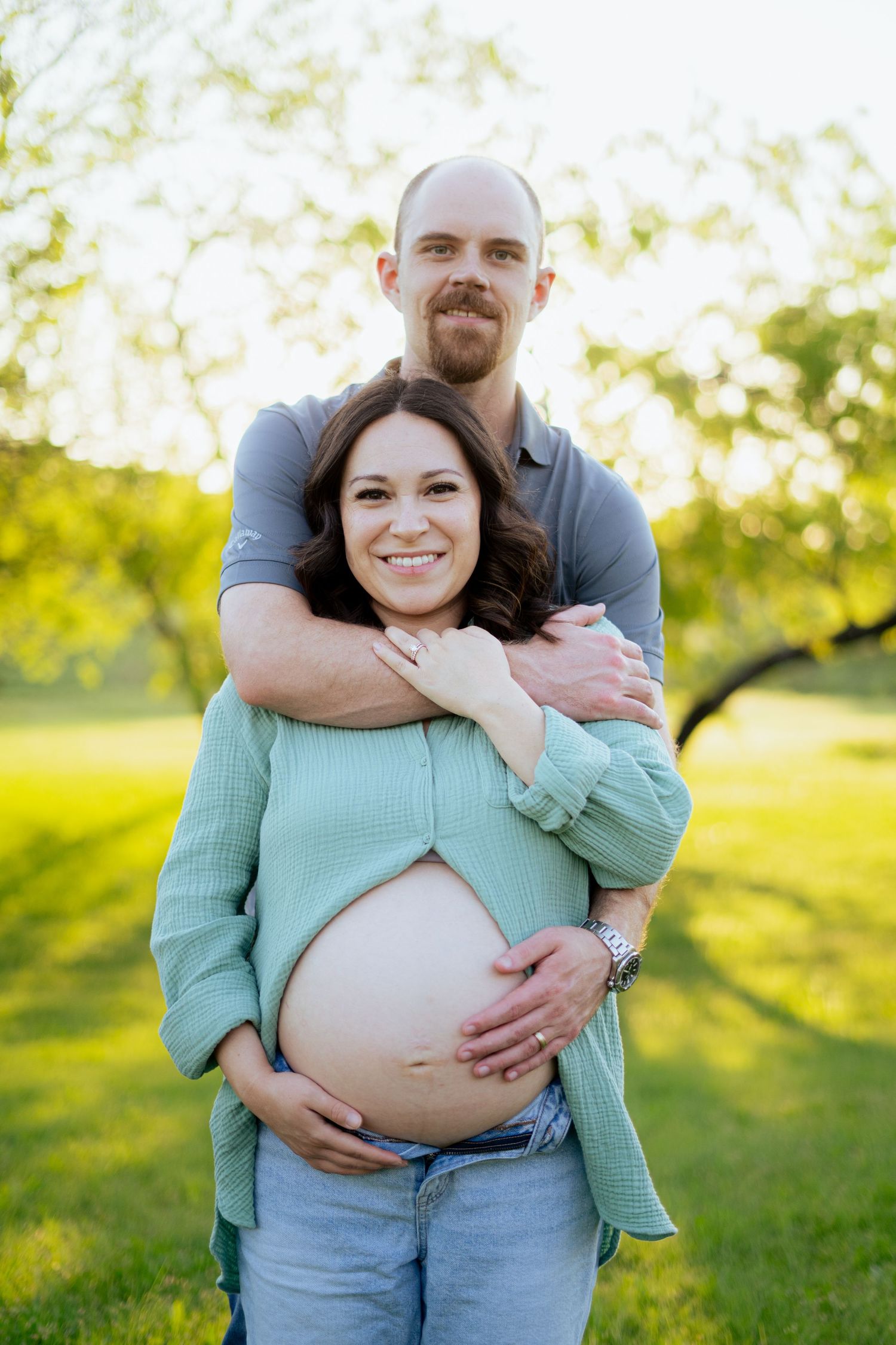 Happy couple embracing outdoors, man hugging pregnant woman from behind while cradling her baby bump in sunlit park.