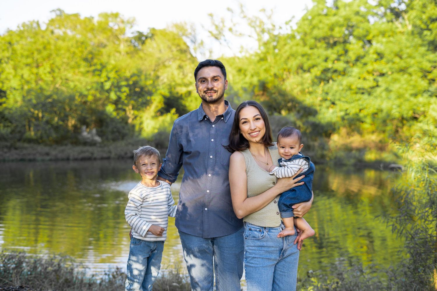 Family of four with two young children pose outdoors near a sunlit pond with green trees.
