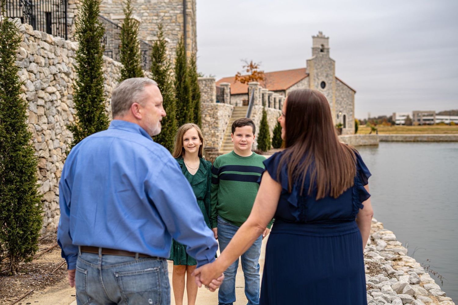 Family of four holding hands and chatting near a stone building by a calm waterfront.
