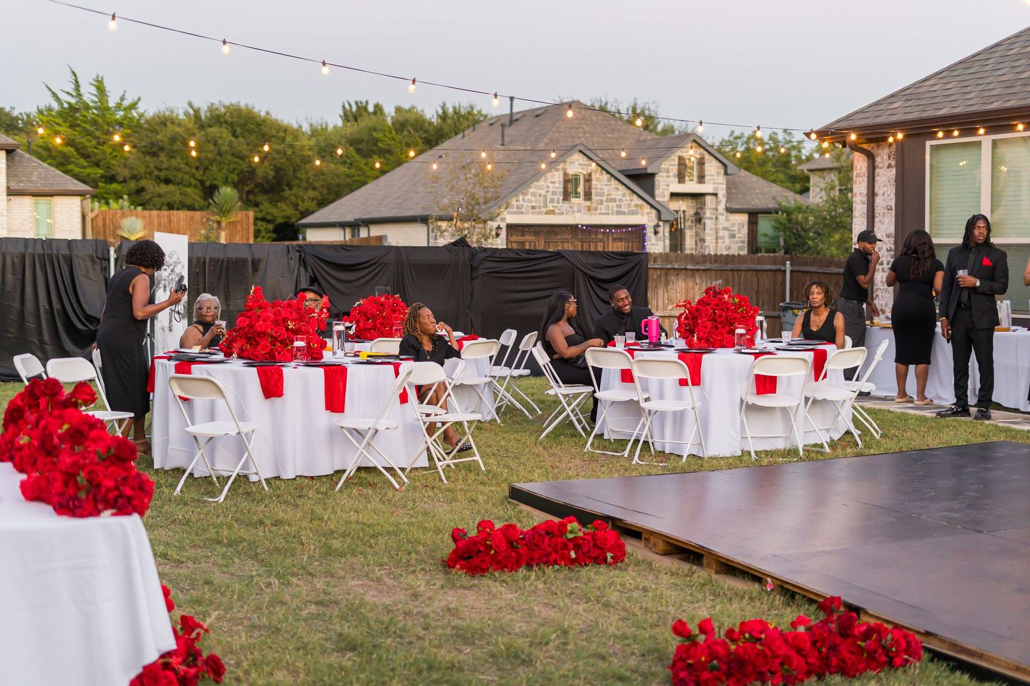 Outdoor backyard wedding reception with white tables, red floral centerpieces, and string lights at dusk.