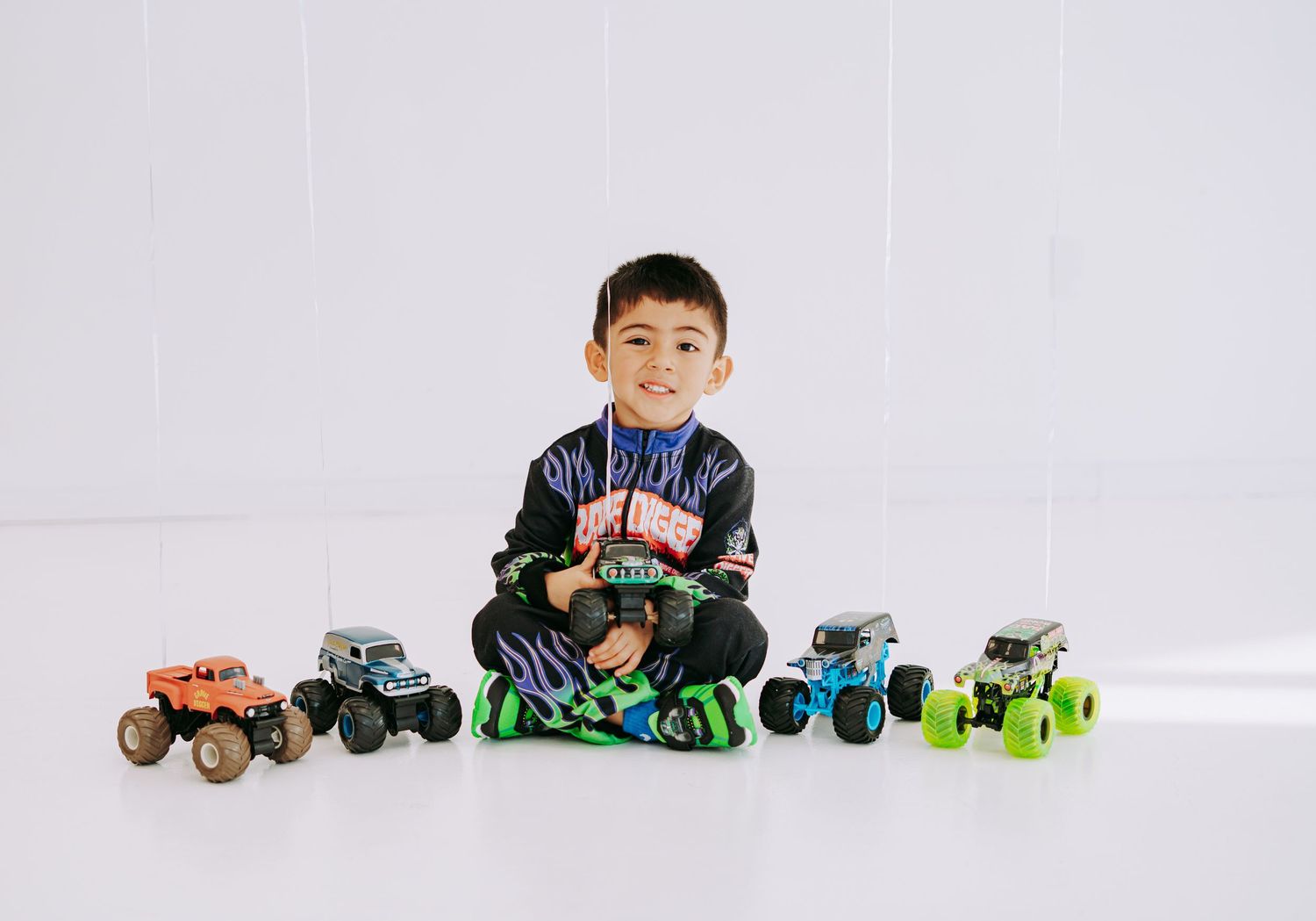 Boy in Monster Jam Grave Digger outfit sitting on floor surrounded by monster truck toys.