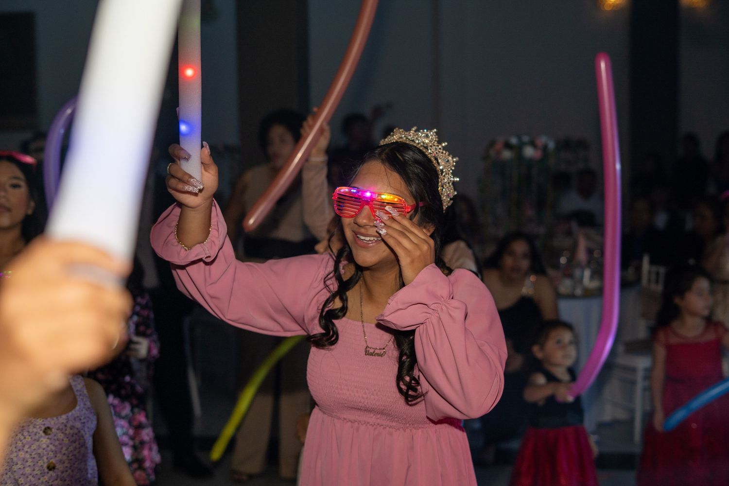Quinceañera honoree in pink dress and tiara dances with glowing neon glasses and light sticks.