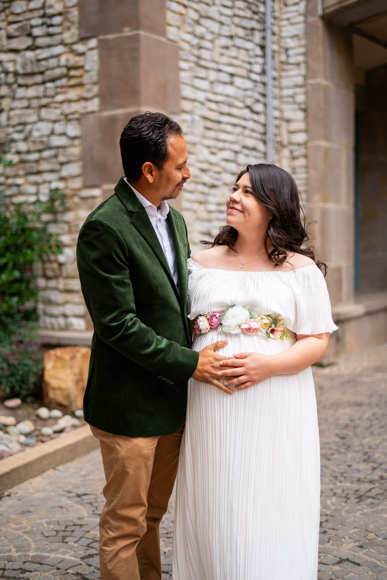 Couple tenderly embracing during maternity photo shoot against rustic stone building backdrop.
