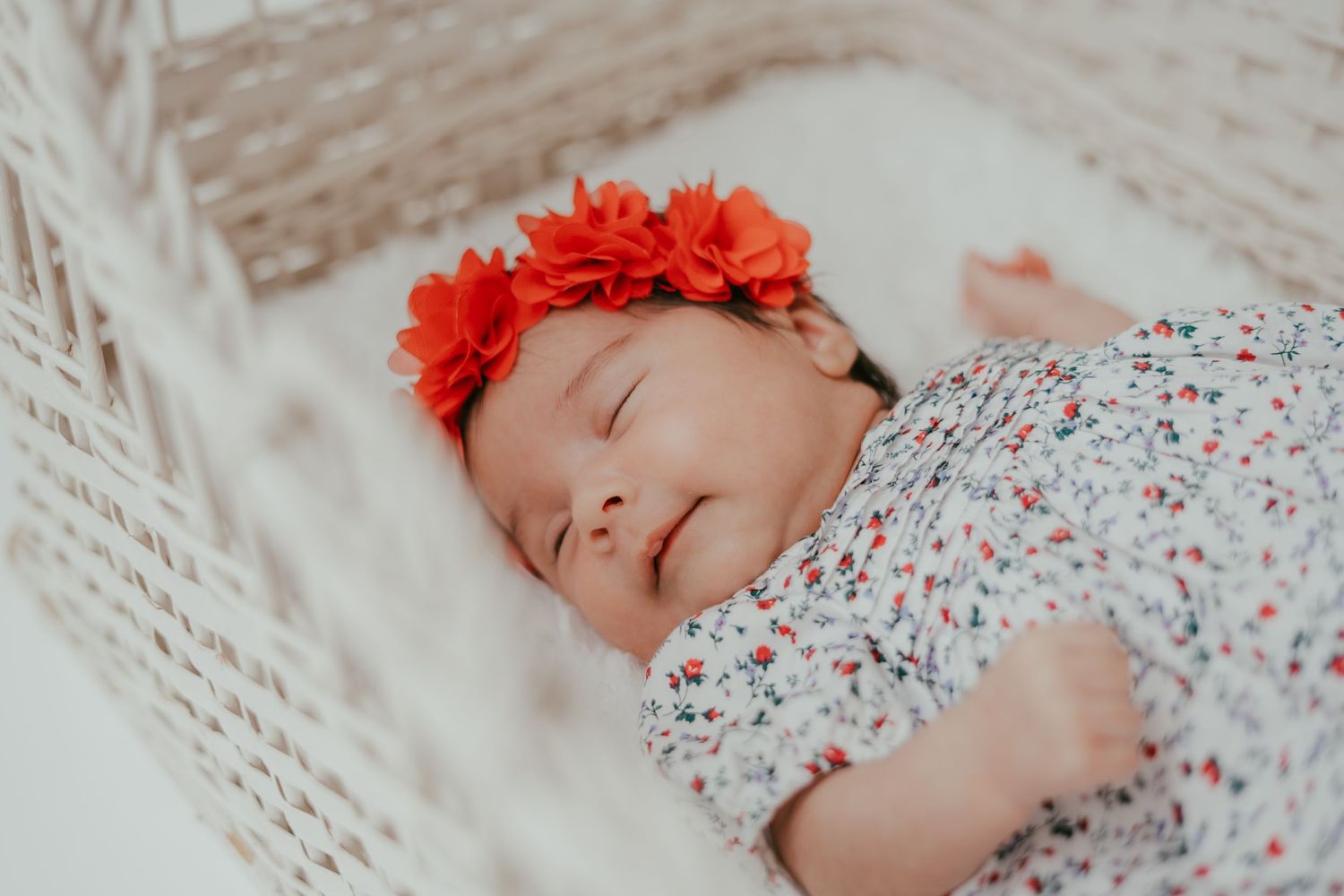 Peaceful newborn baby girl wearing red floral headband sleeps with a smile in a wicker bassinet.