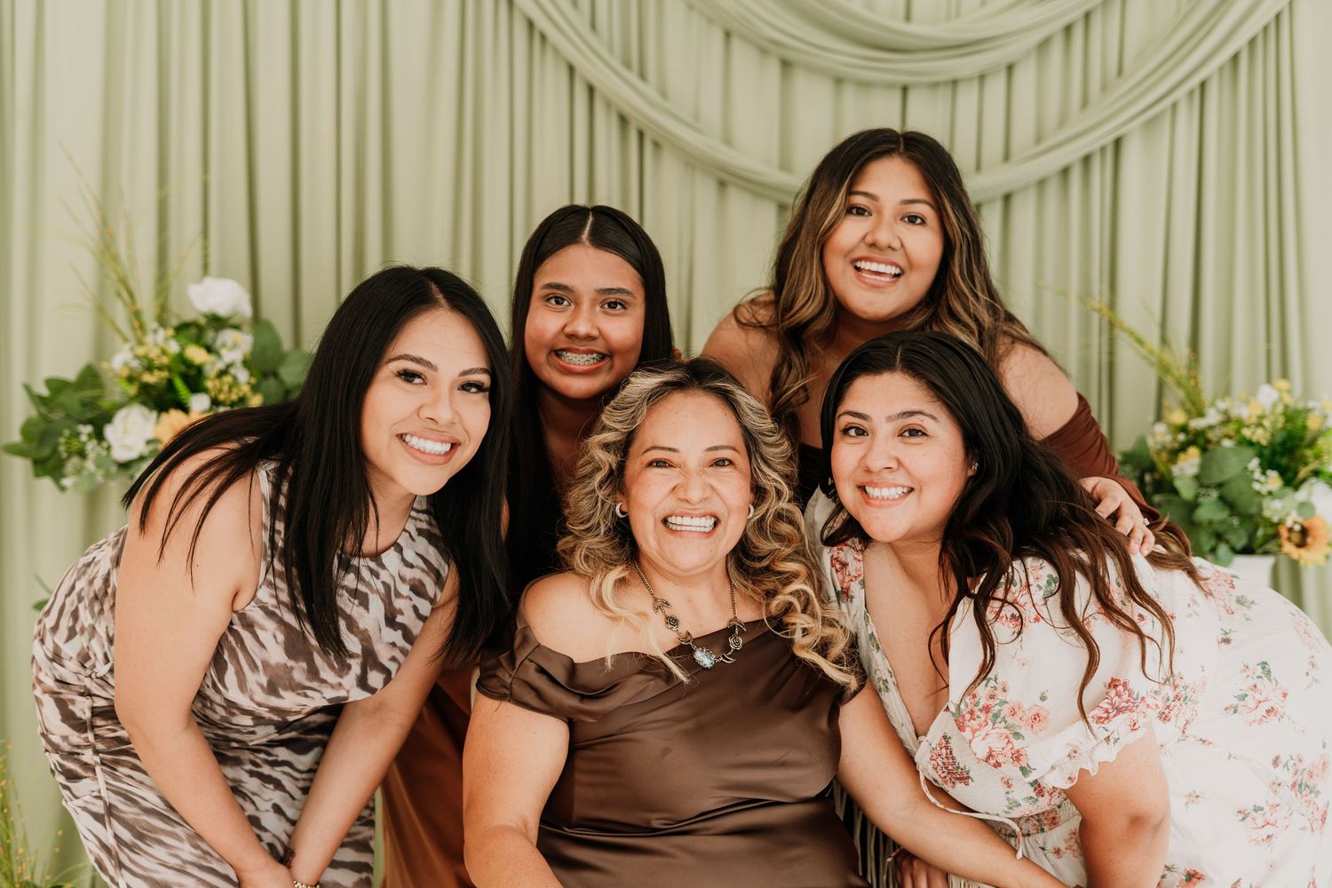 Five women smiling together in front of green floral backdrop at celebration.