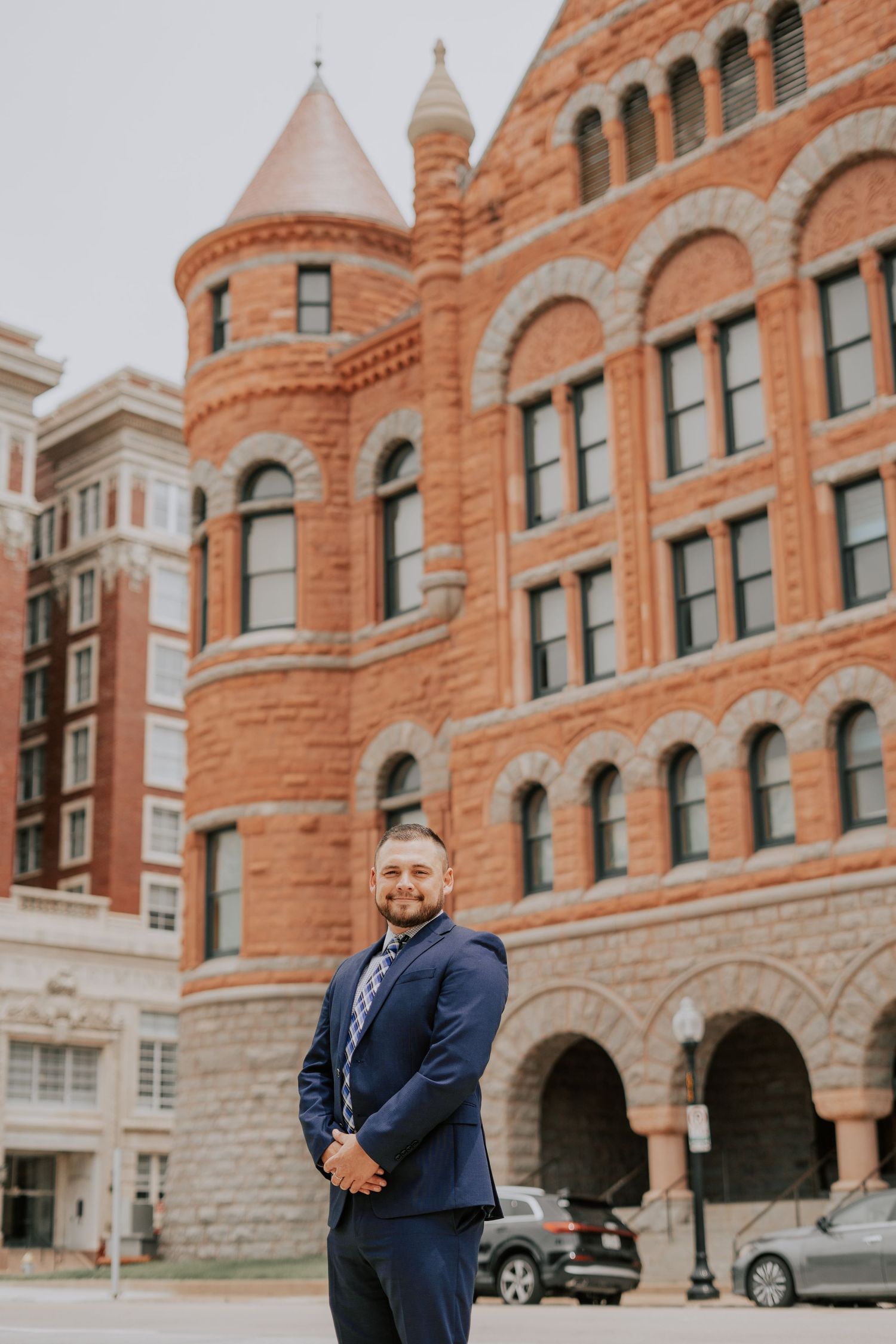Smiling man in navy suit standing in front of historic red brick building with tower.