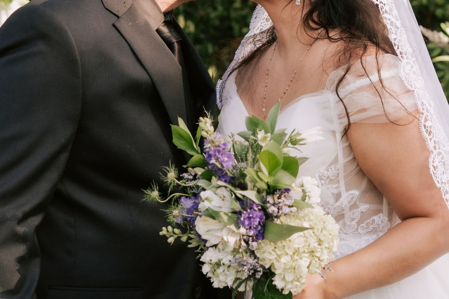 Bride in white gown holding purple and white floral bouquet standing beside groom in black suit outdoors.