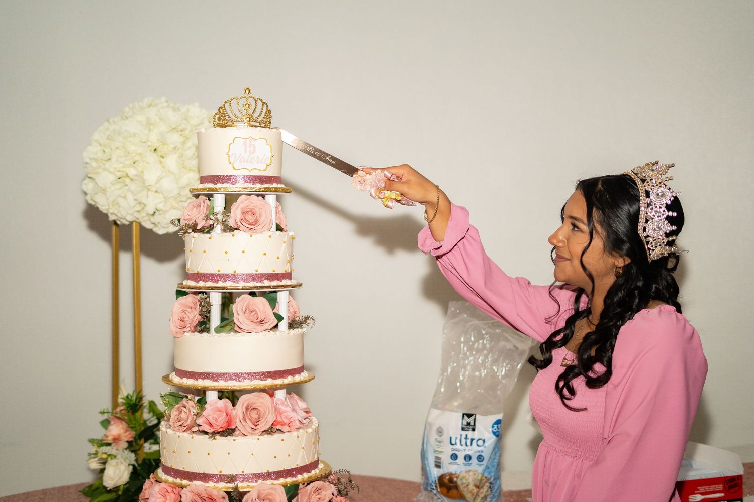 Girl in pink dress and tiara cuts elegant multi-tiered quinceañera cake decorated with pink roses.